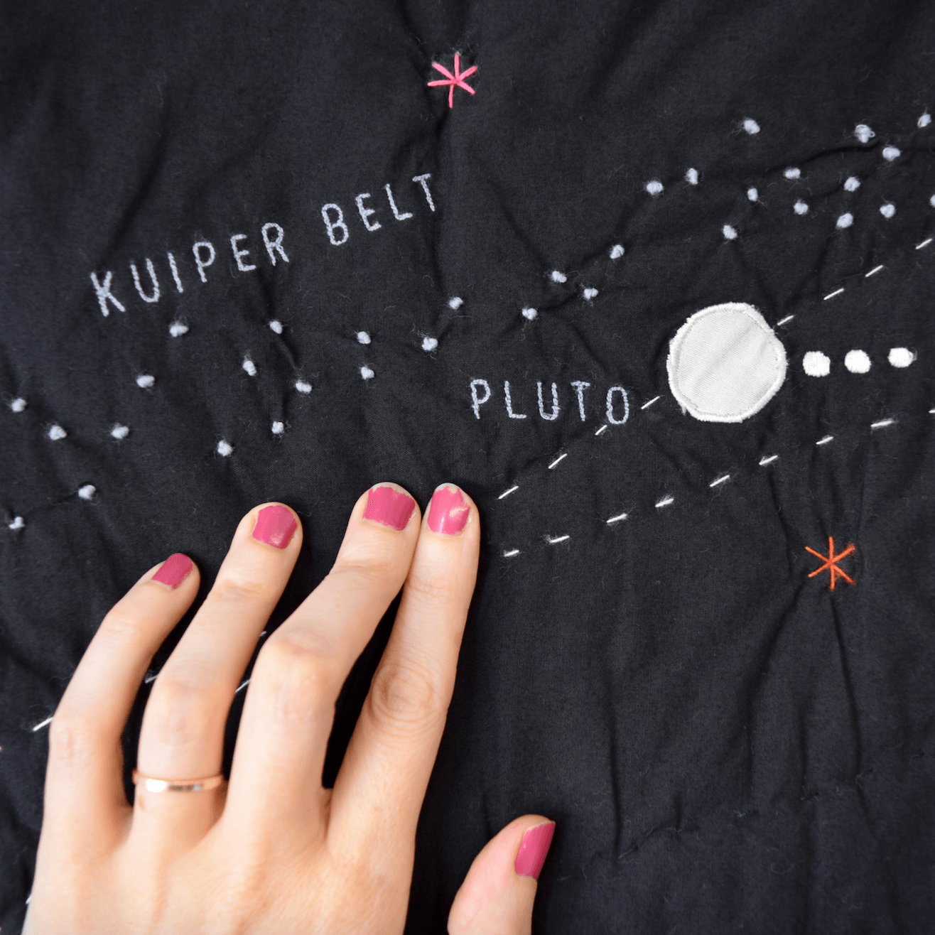 Black fabric with embroidered solar system design, including 'Kuiper Belt' and 'Pluto', with a hand touching it.