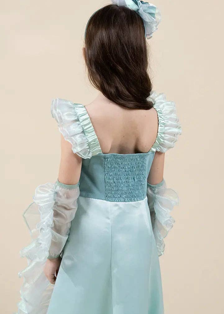 A young girl viewed from behind wearing a light blue and white dress with ruffled off-the-shoulder sleeves, sheer long gloves, and a matching hair bow against a plain beige background.