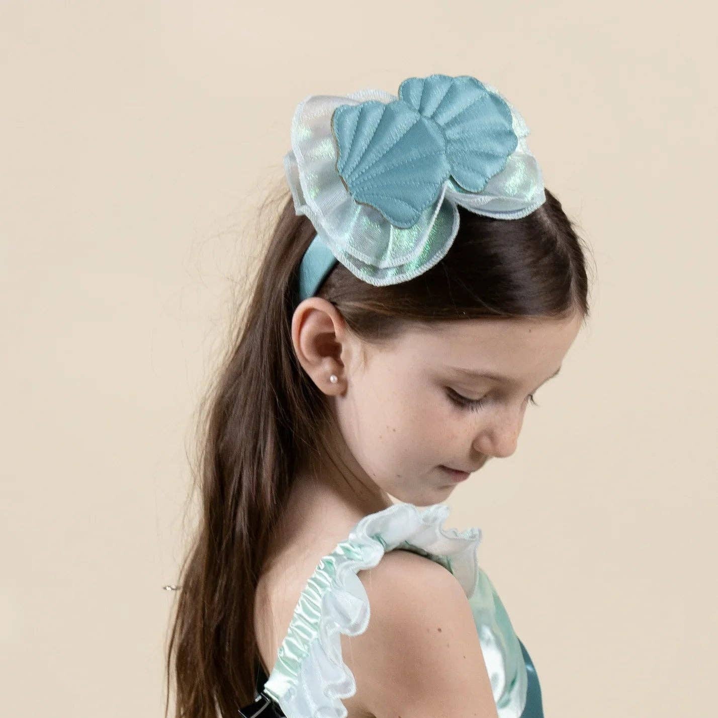 A young girl with long brown hair is shown from the side and back, wearing a light blue costume dress with ruffled trim, sheer white arm cuffs, and a matching light blue shell-shaped hair bow headband against a solid beige background.