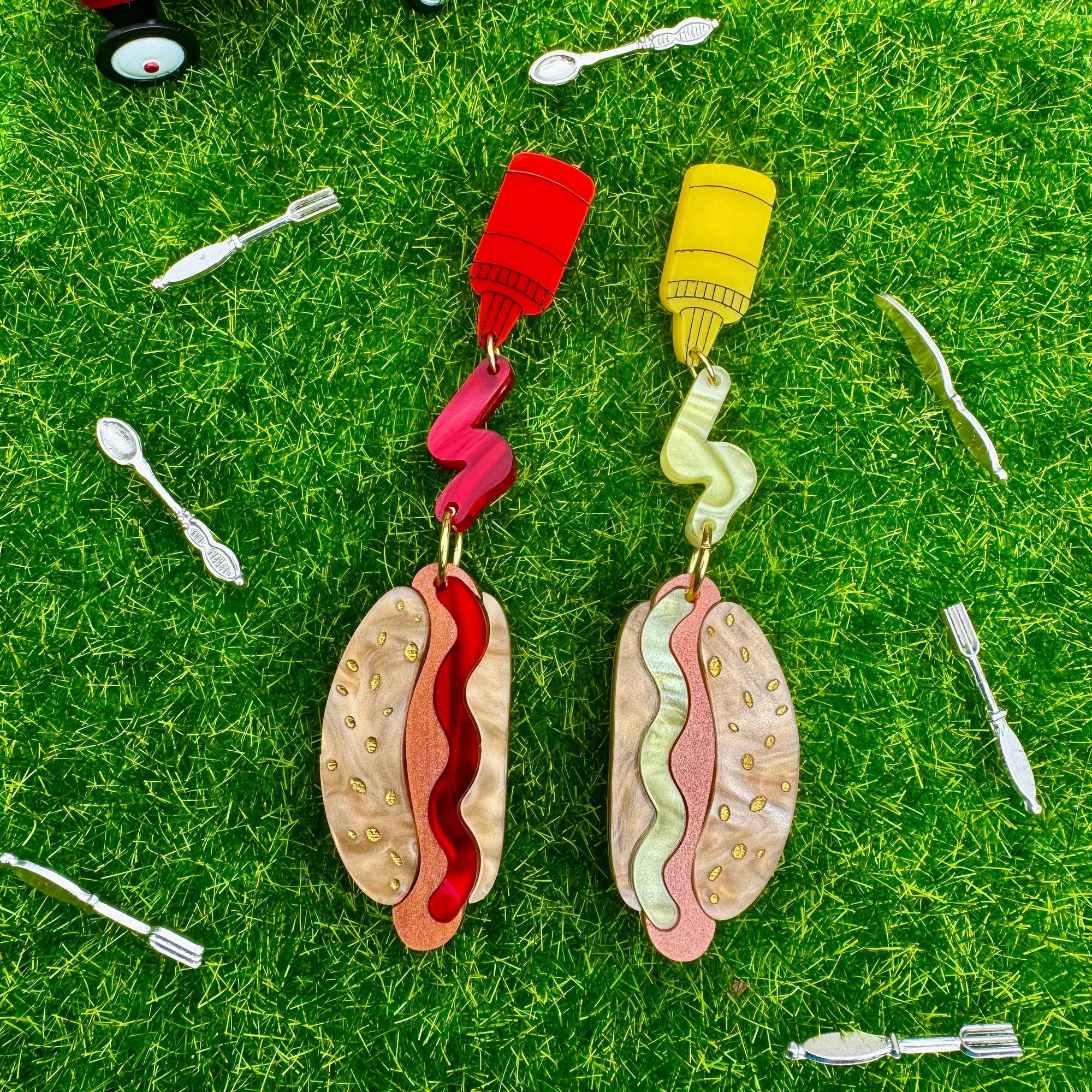 Hot dog and mustard clip-on earrings on grass with a toy wagon and utensils.