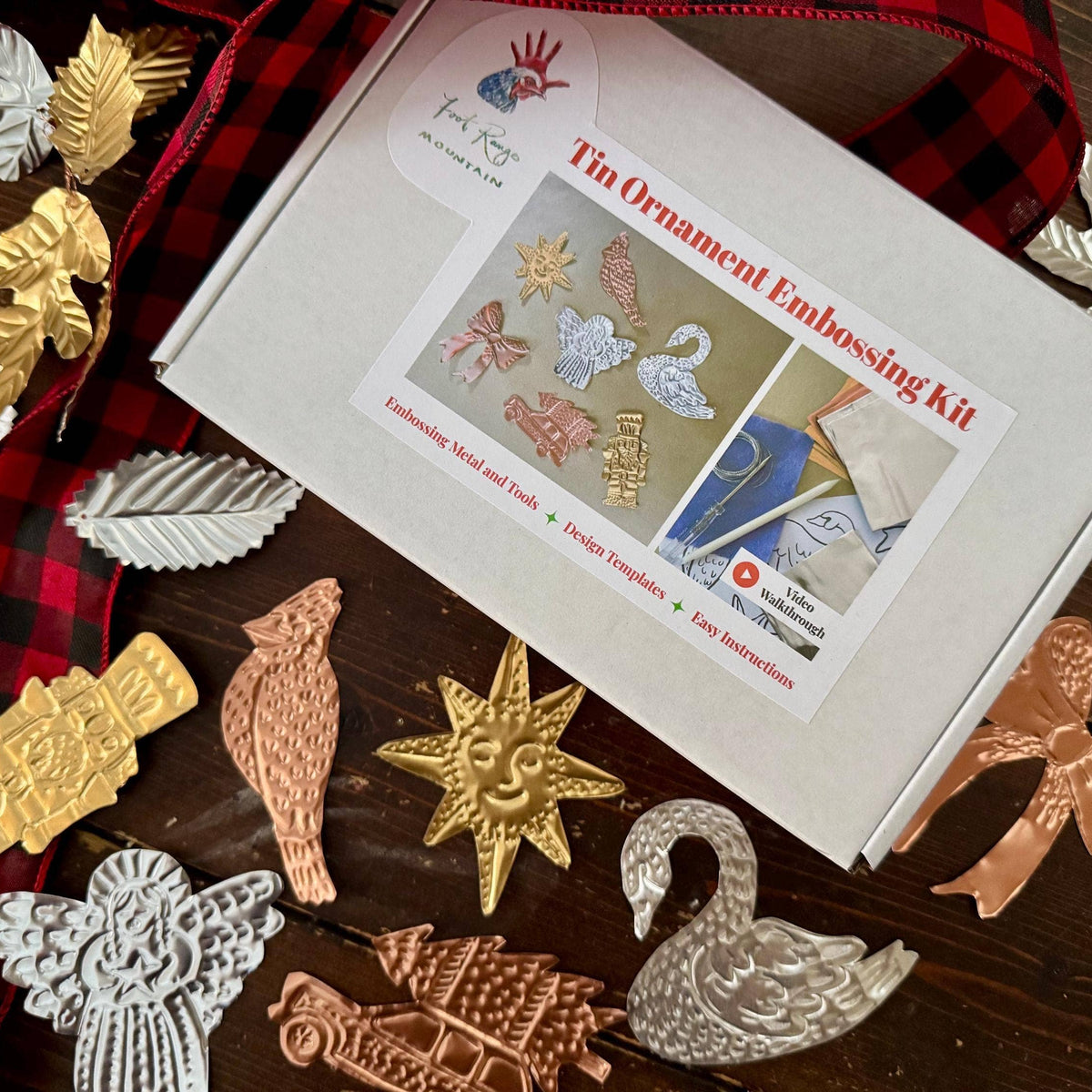 A tin ornament embossing kit box is displayed on a dark wooden surface with a red and black buffalo plaid ribbon. Surrounding the box are various finished metal ornament cutouts in gold, silver, and rose gold, including a swan, sun, bird, and angel.