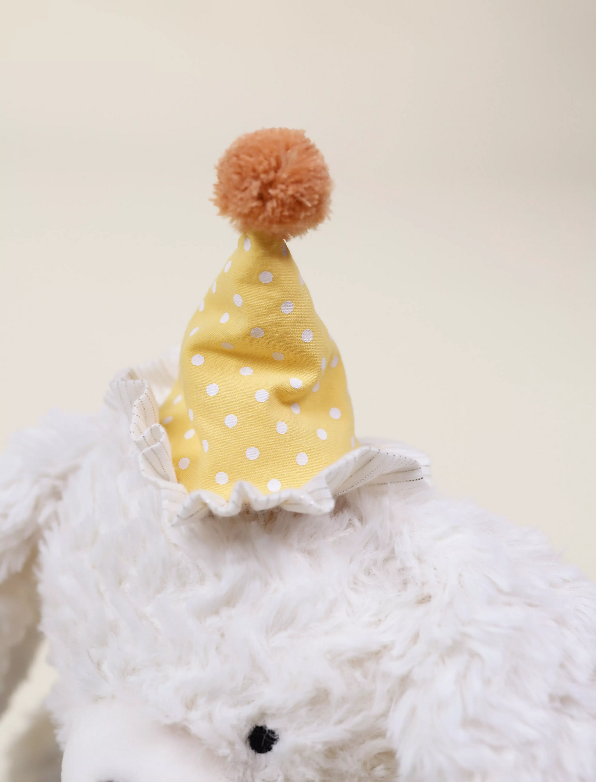 White plush toy wearing a yellow polka dot hat with an orangs pom-pom on a light beige background