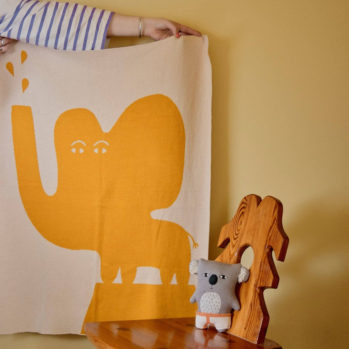 A person holds up a cream-colored woven textile featuring a large mustard yellow elephant graphic, displayed in a nursery-like setting with a wooden chair and a small koala toy against a warm yellow wall.