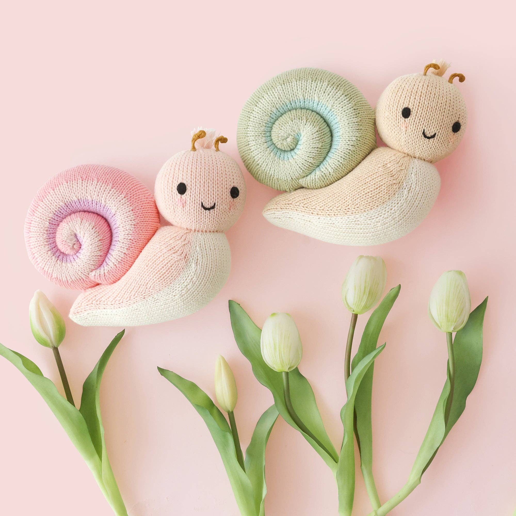 Three soft toy snails with colorful shells on a pink background with tulips.