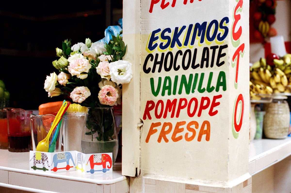 A close-up of a white wooden sign with colorful handwritten text reading 'ESKIMOS CHOCOLATE VAINILLA ROMPOPE FRESA' in red, blue, green, and orange. A vase of pink and white flowers, drink cups, and other cafe items are visible on the counter beside it.
