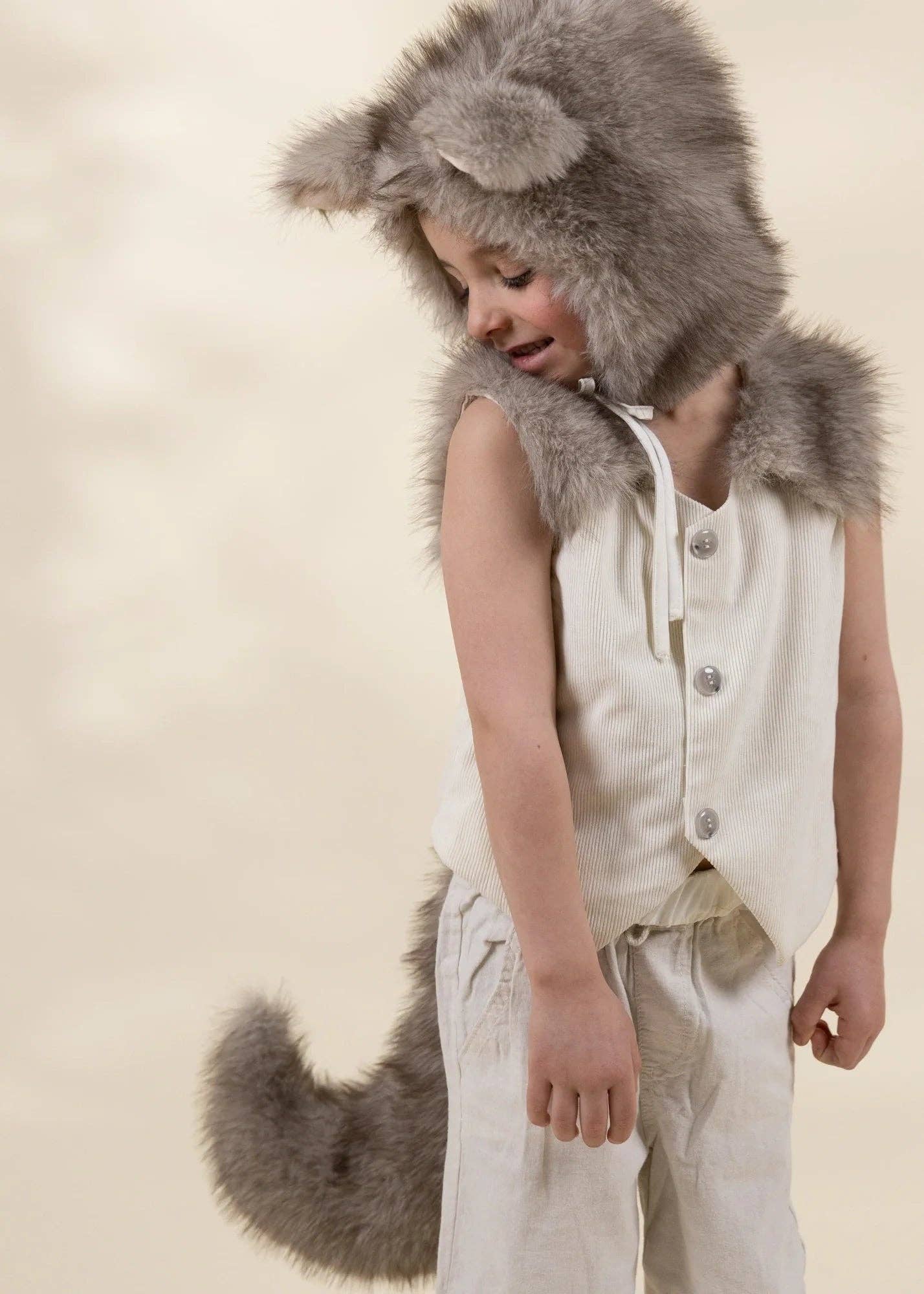 A child wearing a wolf costume with a gray faux-fur hood featuring wolf ears, a matching gray tail, and a cream linen vest and pants, posed against a warm beige background.