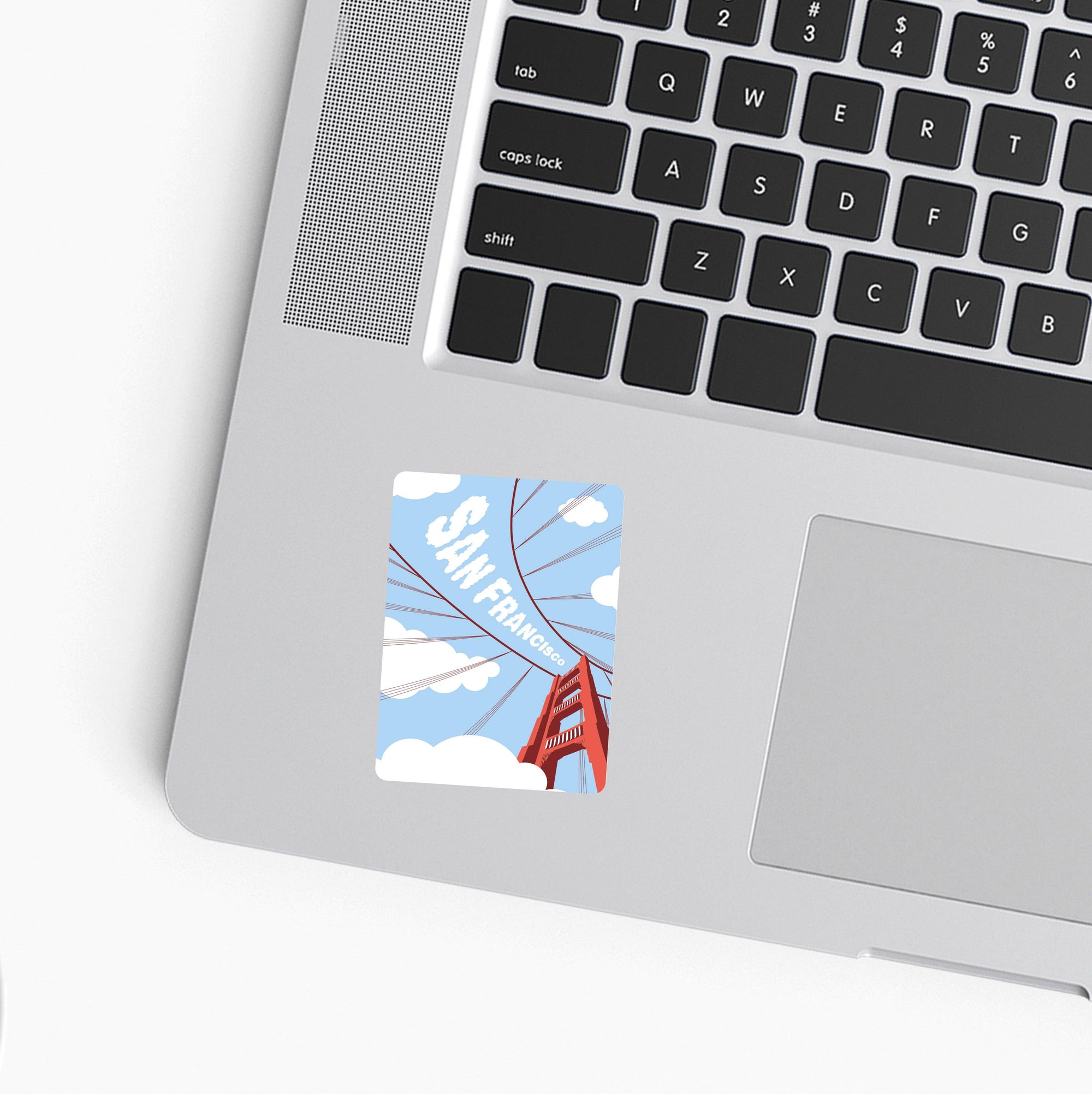 A square sticker featuring a stylized illustration of the Golden Gate Bridge in San Francisco under a blue sky, shown applied to the palm rest of a silver laptop.