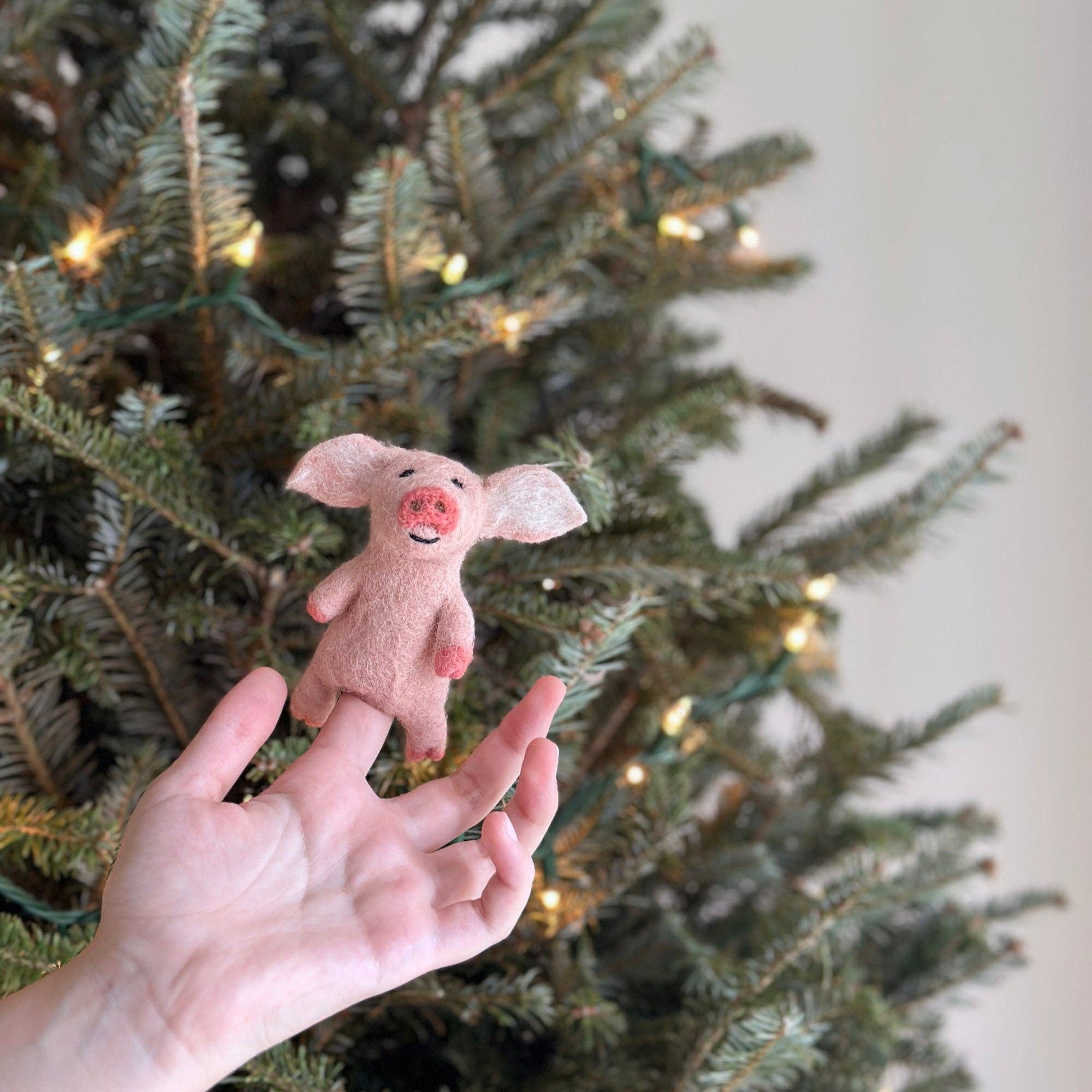Small pink pig plush toy held in front of a decorated Christmas tree.