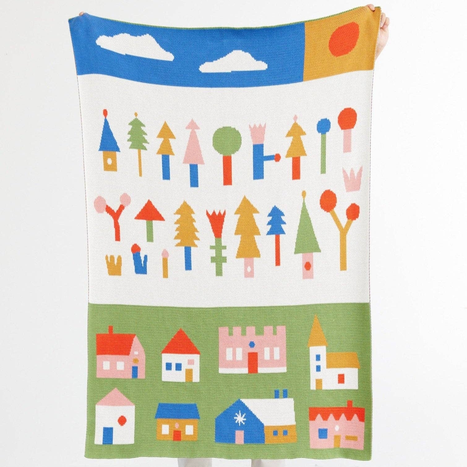 Colorful children's blanket with houses, trees, and a sun design held by a person on a white background.