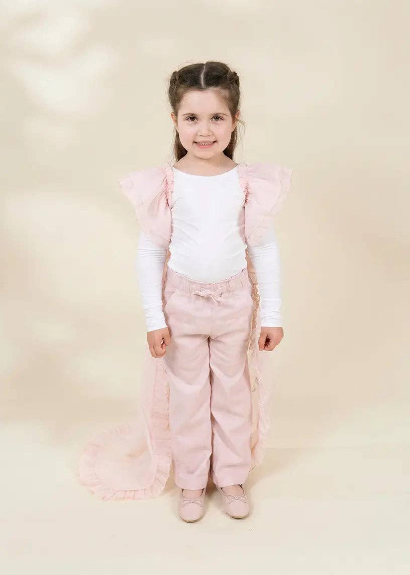 A young girl stands against a warm beige studio background wearing a white long-sleeve top, light pink pants, and a matching sheer pink ruffled cape with a long train.