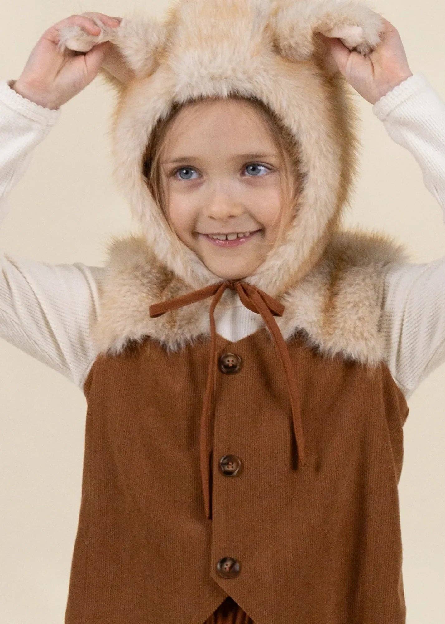 A young child wearing a brown faux-suede vest with a fluffy beige faux-fur hood and matching tie, over a cream ribbed long-sleeve top, against a plain beige background.