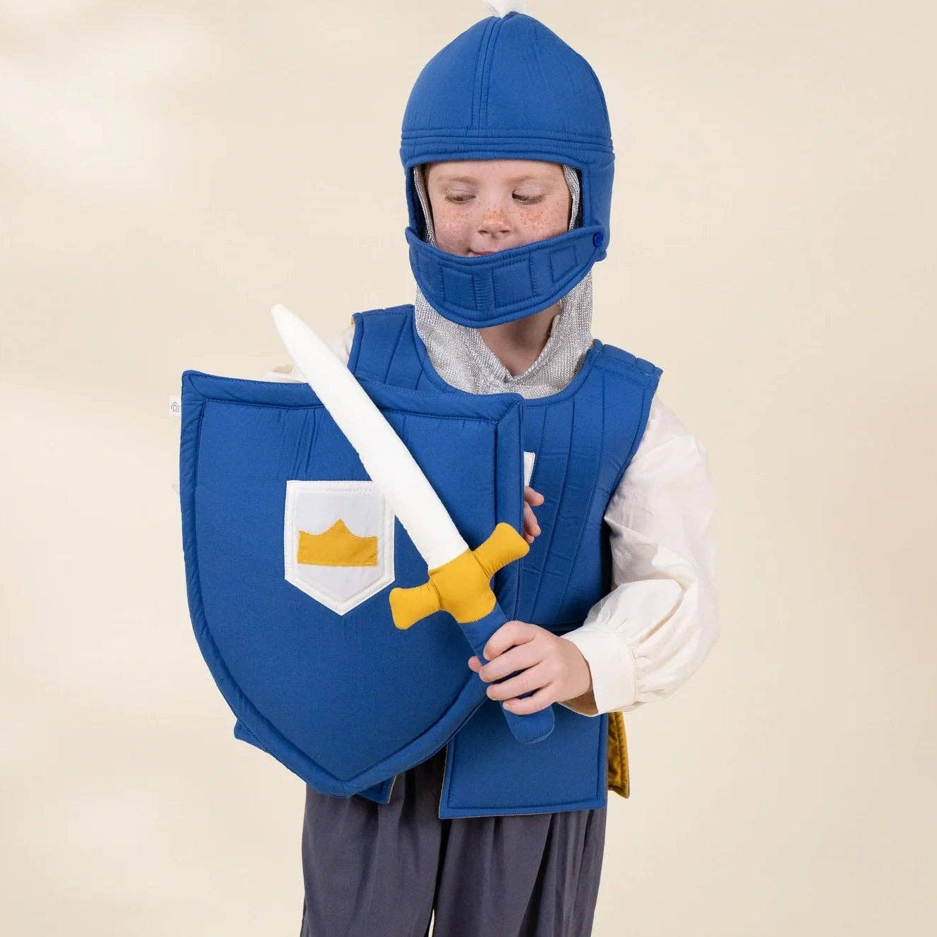 A child wearing a medieval knight costume, including a blue helmet, blue padded vest, and a blue shield with a white crest and gold crown, holding a toy sword with a yellow hilt, against a plain beige background.