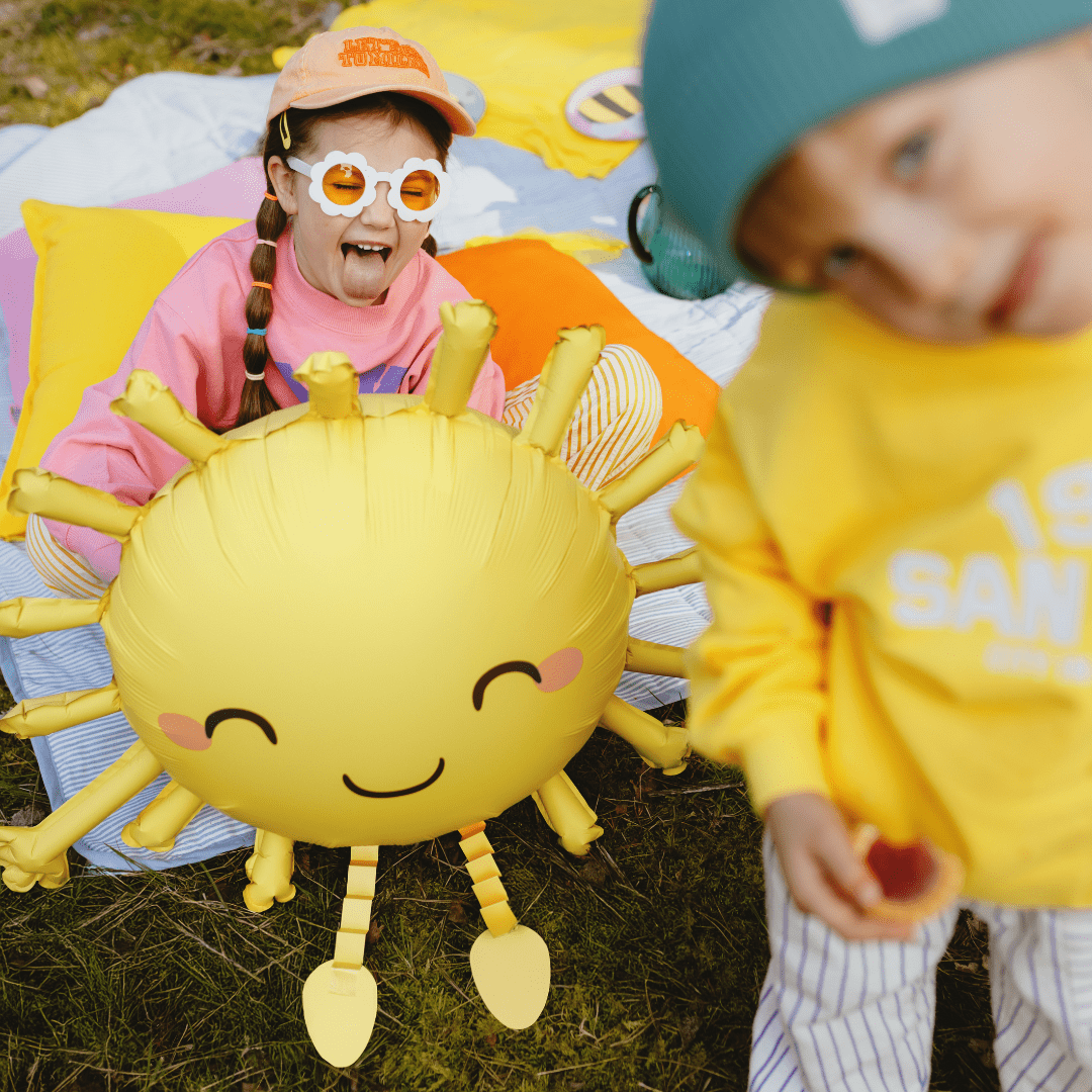 A child wearing a pink sweatshirt and orange cap sits on a colorful blanket outdoors, smiling while wearing novelty white sunglasses with orange lenses. In the foreground, another child in a yellow sweatshirt and teal beanie stands out of focus.