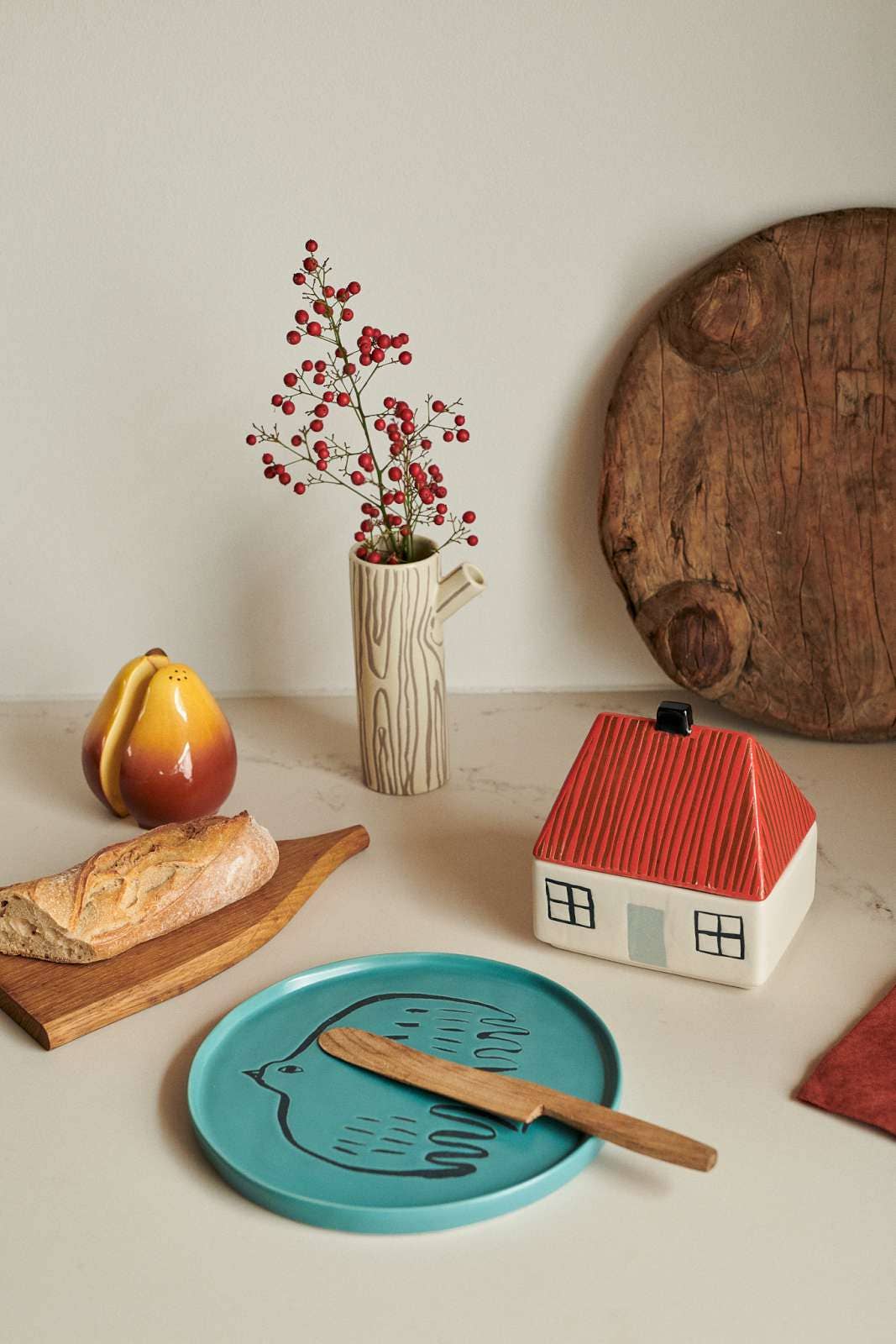 A styled tabletop scene with a turquoise plate holding a wooden knife, next to a small house-shaped container with a red roof, a vase with red berries, bread on a wooden board, and a pear against a warm neutral background.
