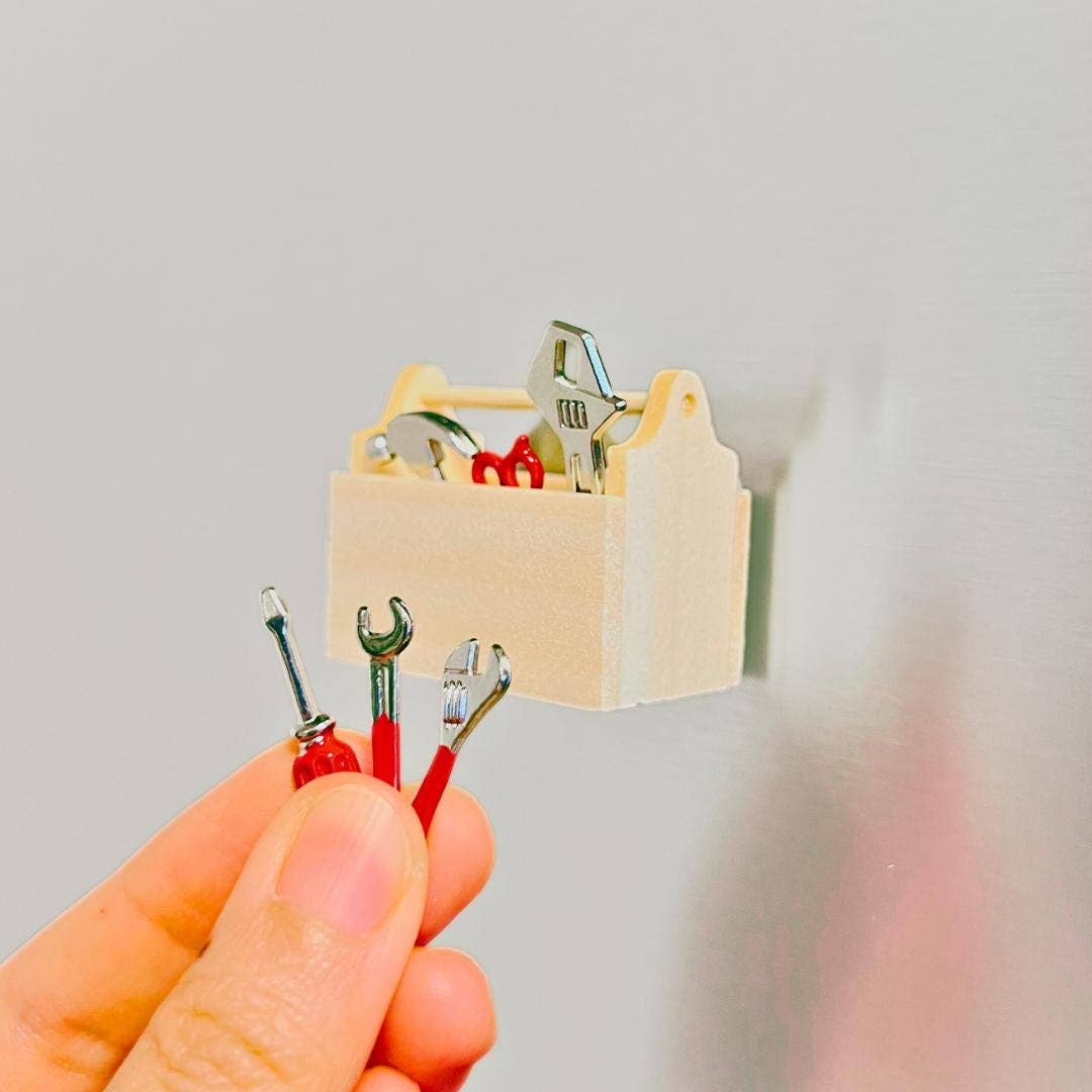 A hand holds miniature wrenches and screwdrivers in front of a small beige wall-mounted tool box filled with tiny tools, against a plain light gray wall.