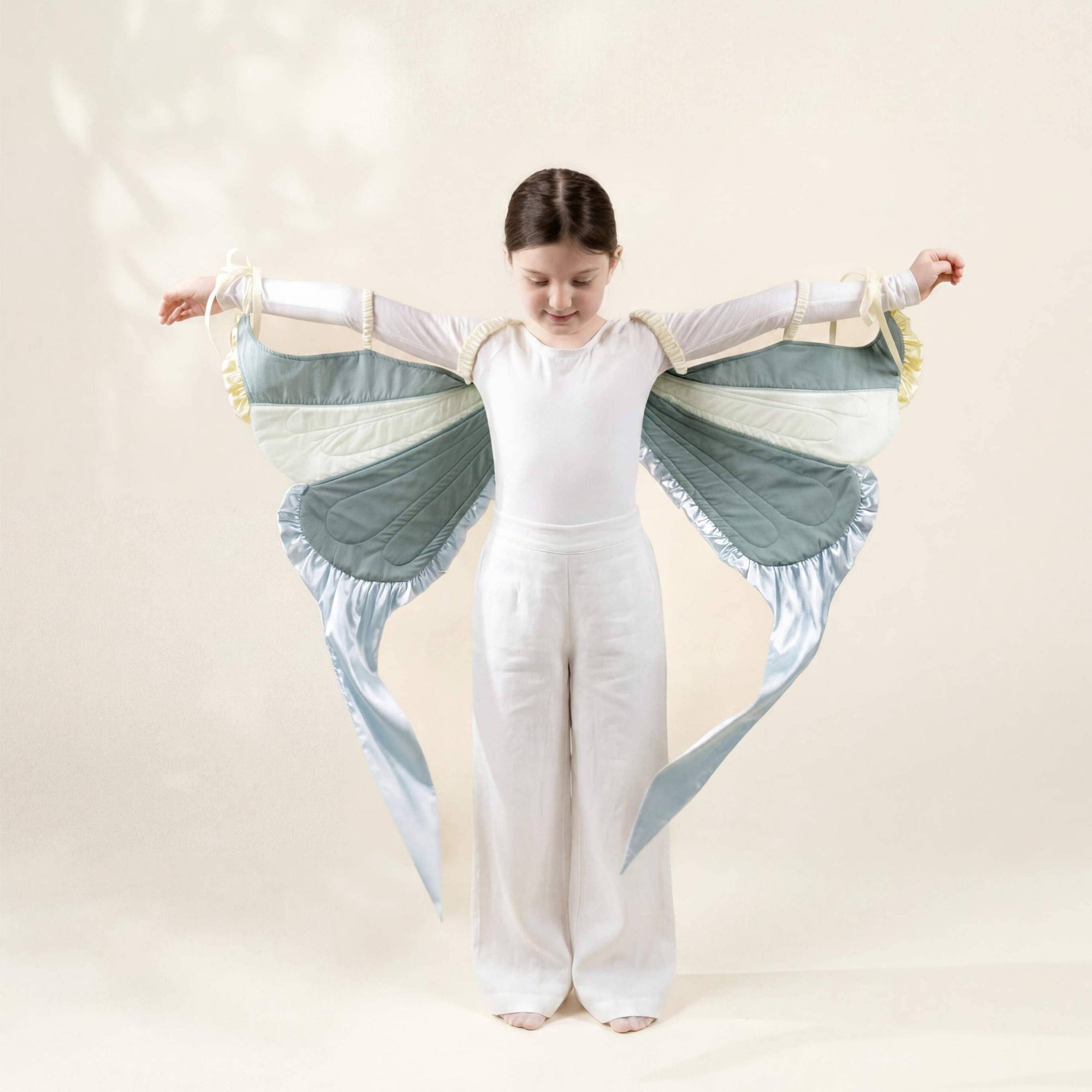 Child wearing a white outfit with green and blue wings against a beige background