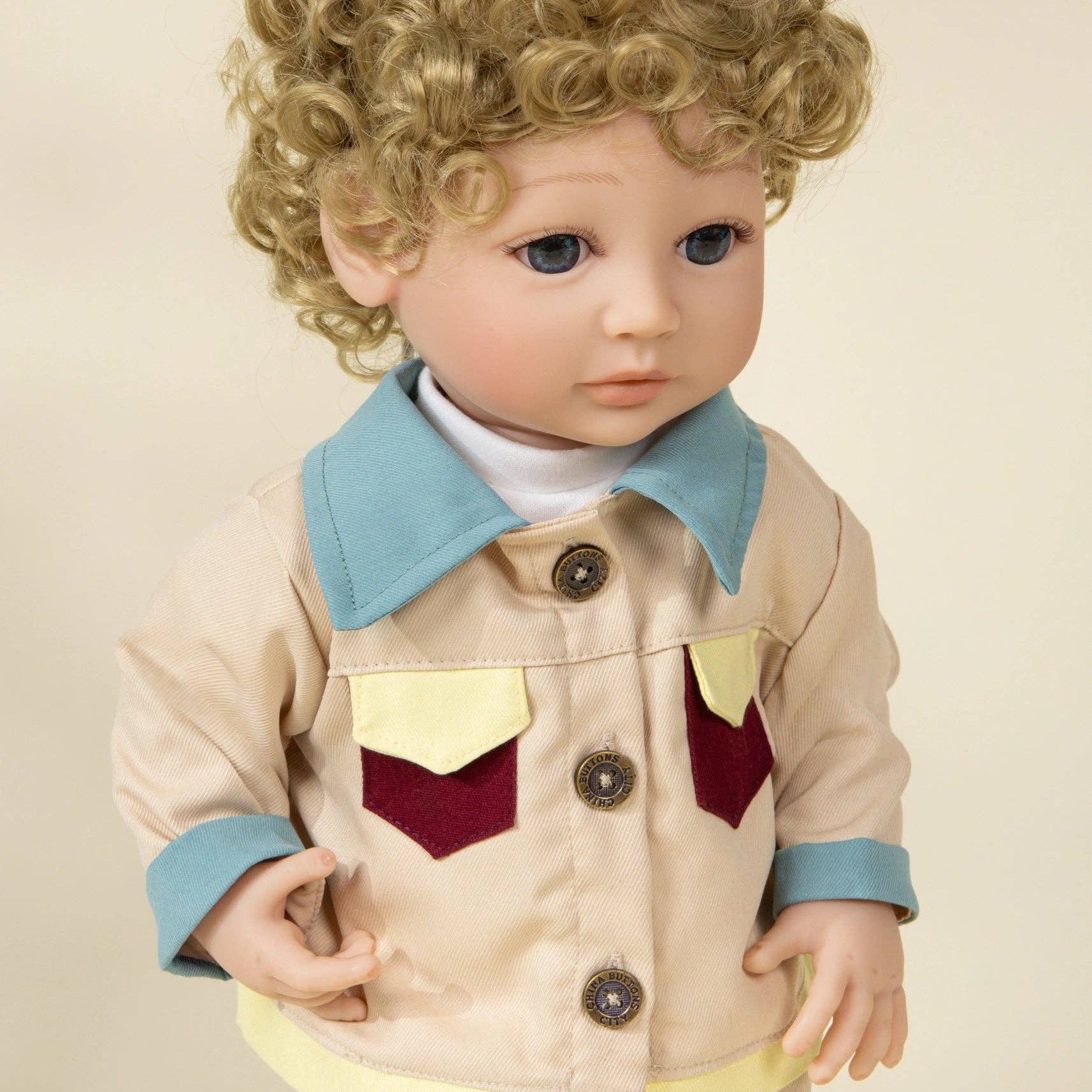Child-sized doll wearing a beige jacket with blue collar and red and yellow accents on a beige background