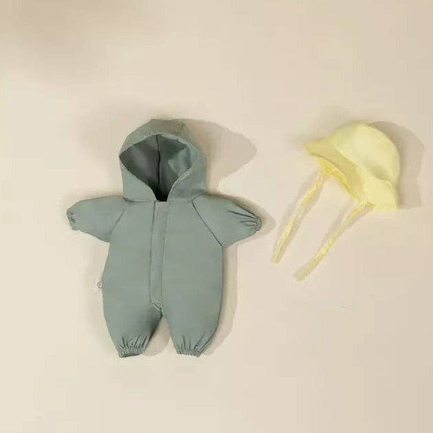 A baby hooded one-piece romper in dusty sage green is laid flat next to a pale yellow bonnet with ties on a warm off-white background.