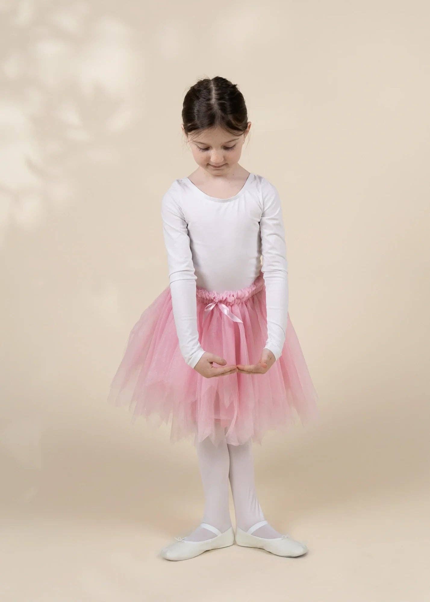 A young girl dancer wearing a white long-sleeve leotard, a pink tulle tutu skirt, white tights, and white ballet slippers, standing against a warm beige background.