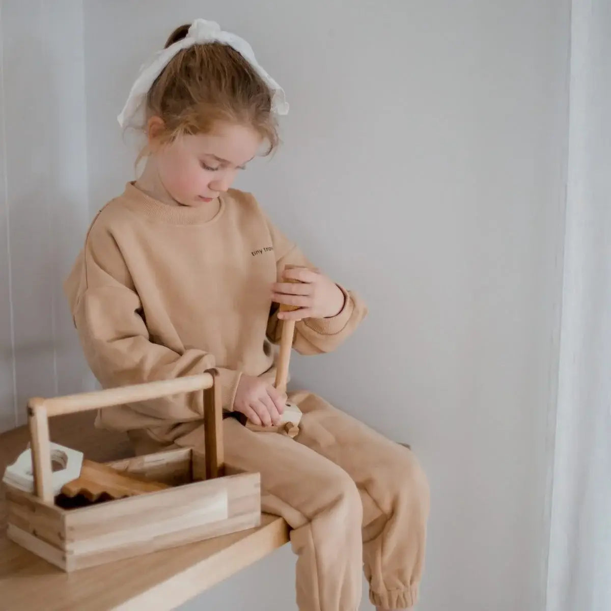 Child in a beige outfit playing with wooden tools in a box.