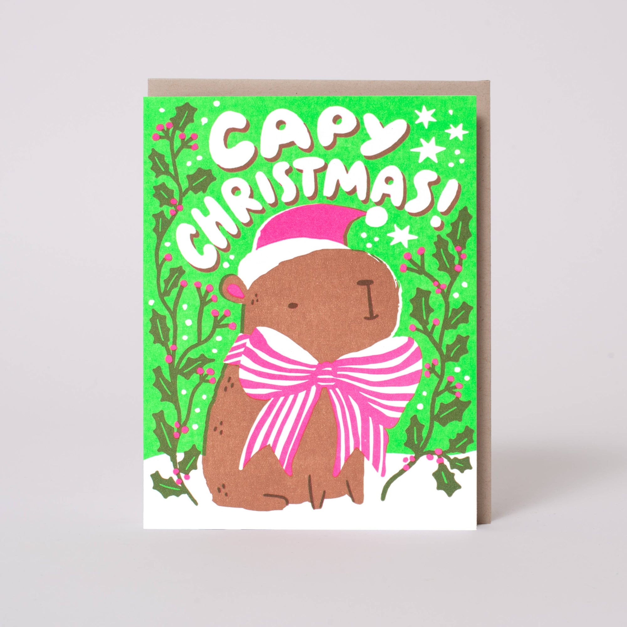 A Christmas greeting card featuring a brown cartoon bear wearing a pink Santa hat and a large pink bow, with the text 'CAPY CHRISTMAS!' on a bright green background with holly and stars. A kraft envelope is partially visible behind the card.