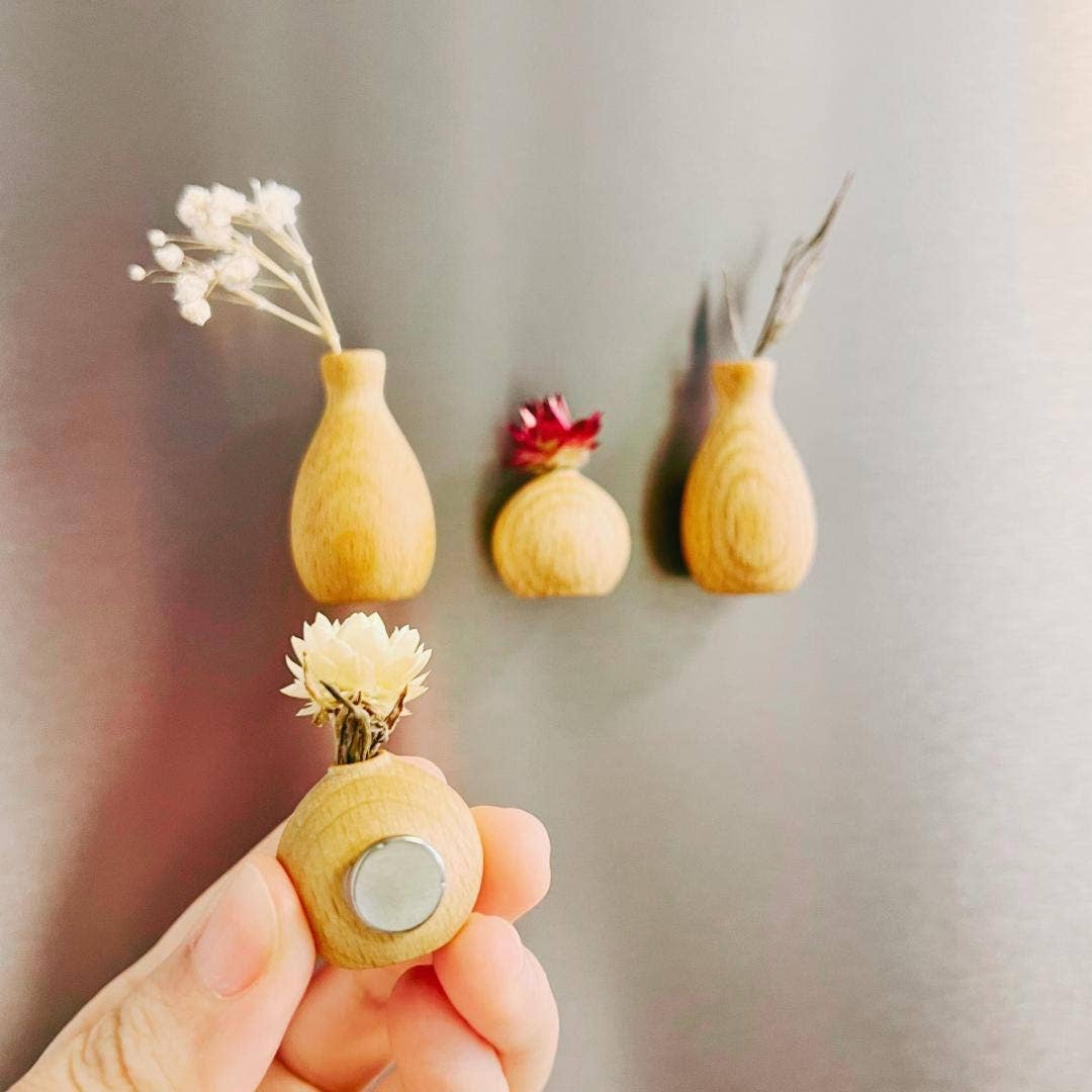 Three small wooden vase-shaped magnets are shown on a gray refrigerator, with one held in a hand in the foreground. The magnets have a natural light wood finish and are decorated with dried white and red flowers.