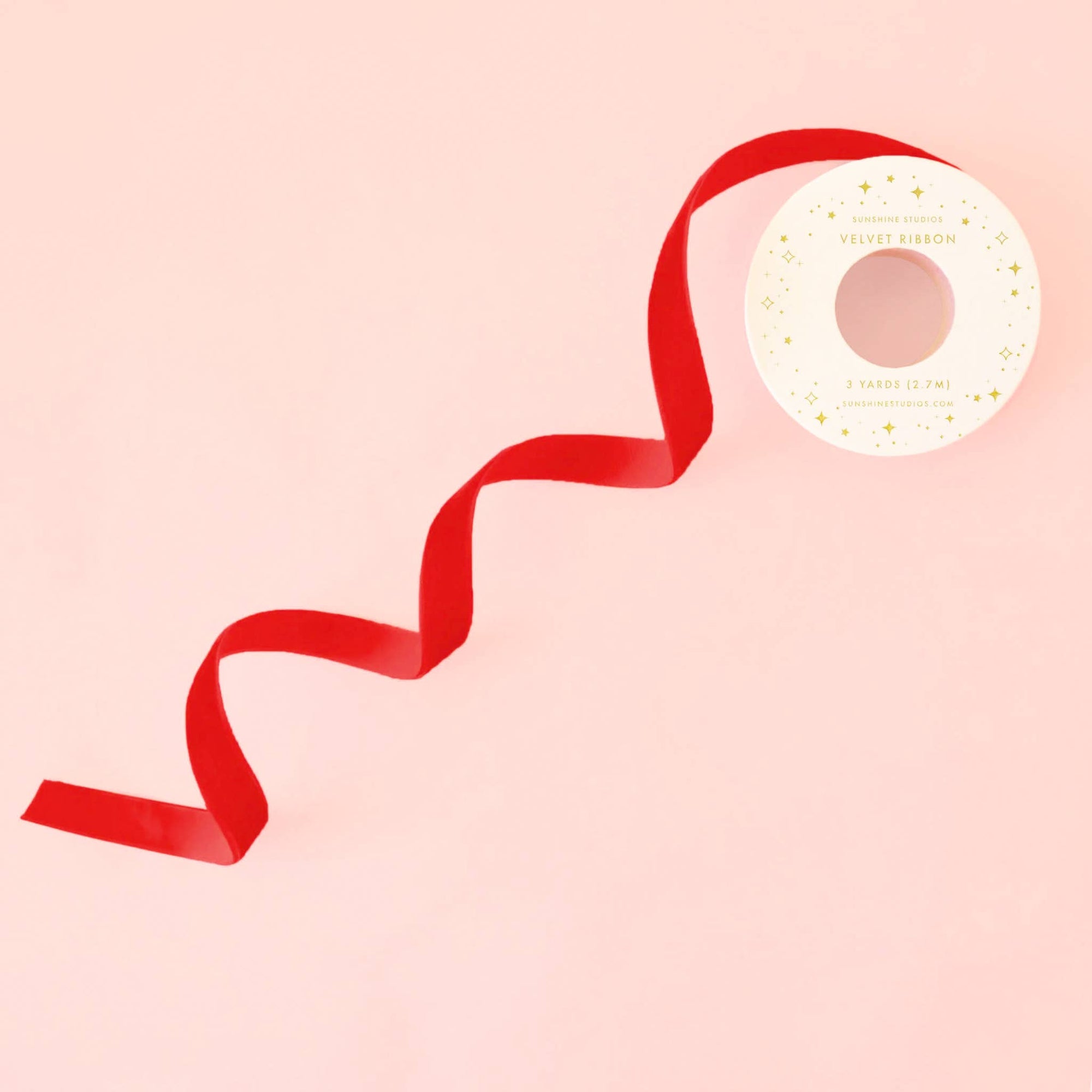 A red velvet ribbon wound on a white spool with gold lettering, set against a solid pale pink background.