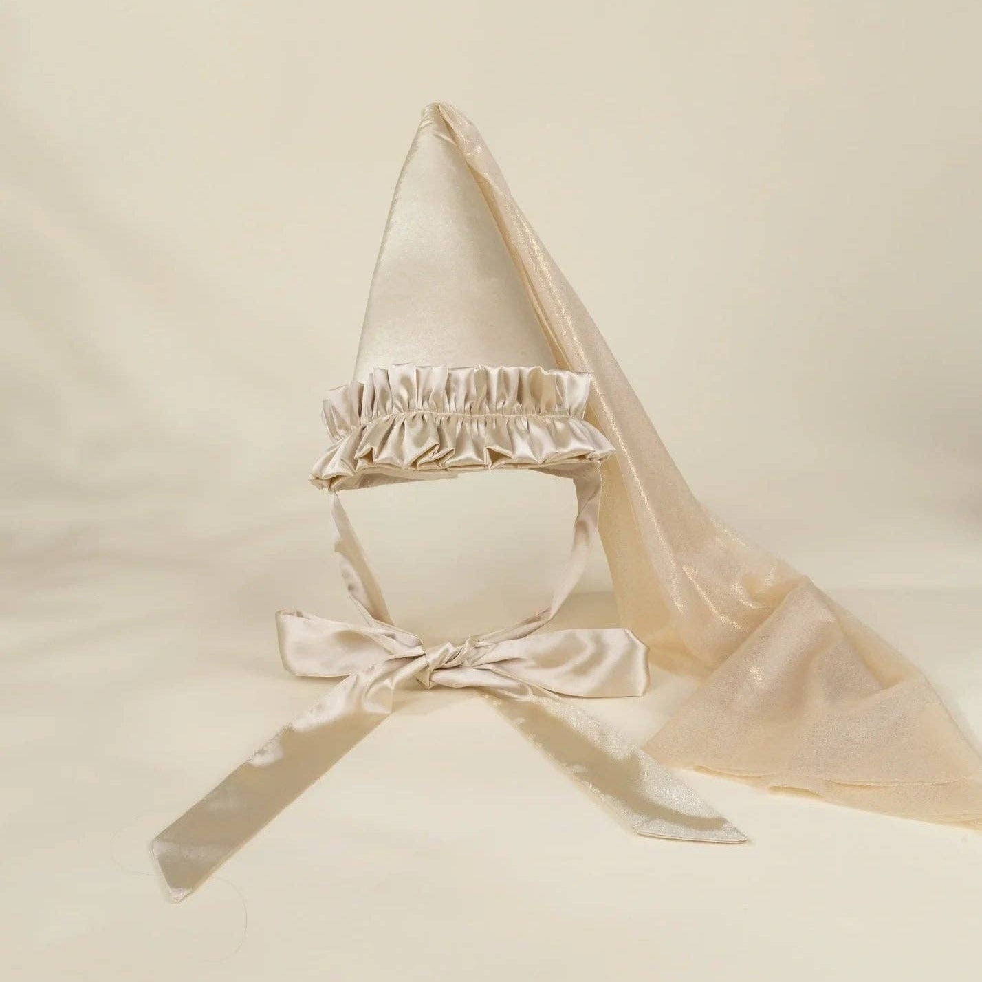 A cream satin baby bonnet with a ruffled trim, chin strap, and attached sheer veil, displayed on a neutral cream background.