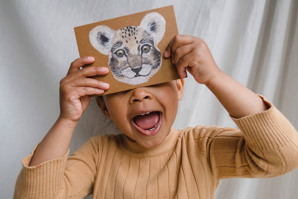 A young child wearing a beige ribbed knit sweater holds a square cardboard card with a painted leopard face over one eye, smiling with their mouth open against a soft white curtain background.