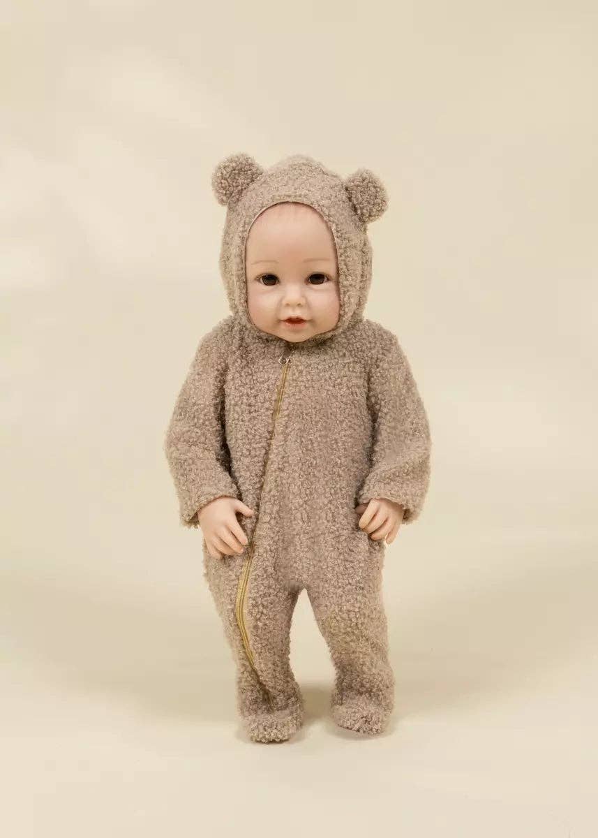 A baby doll wearing a fuzzy teddy bear one-piece outfit with a hood featuring bear ears, standing against a plain beige studio background.