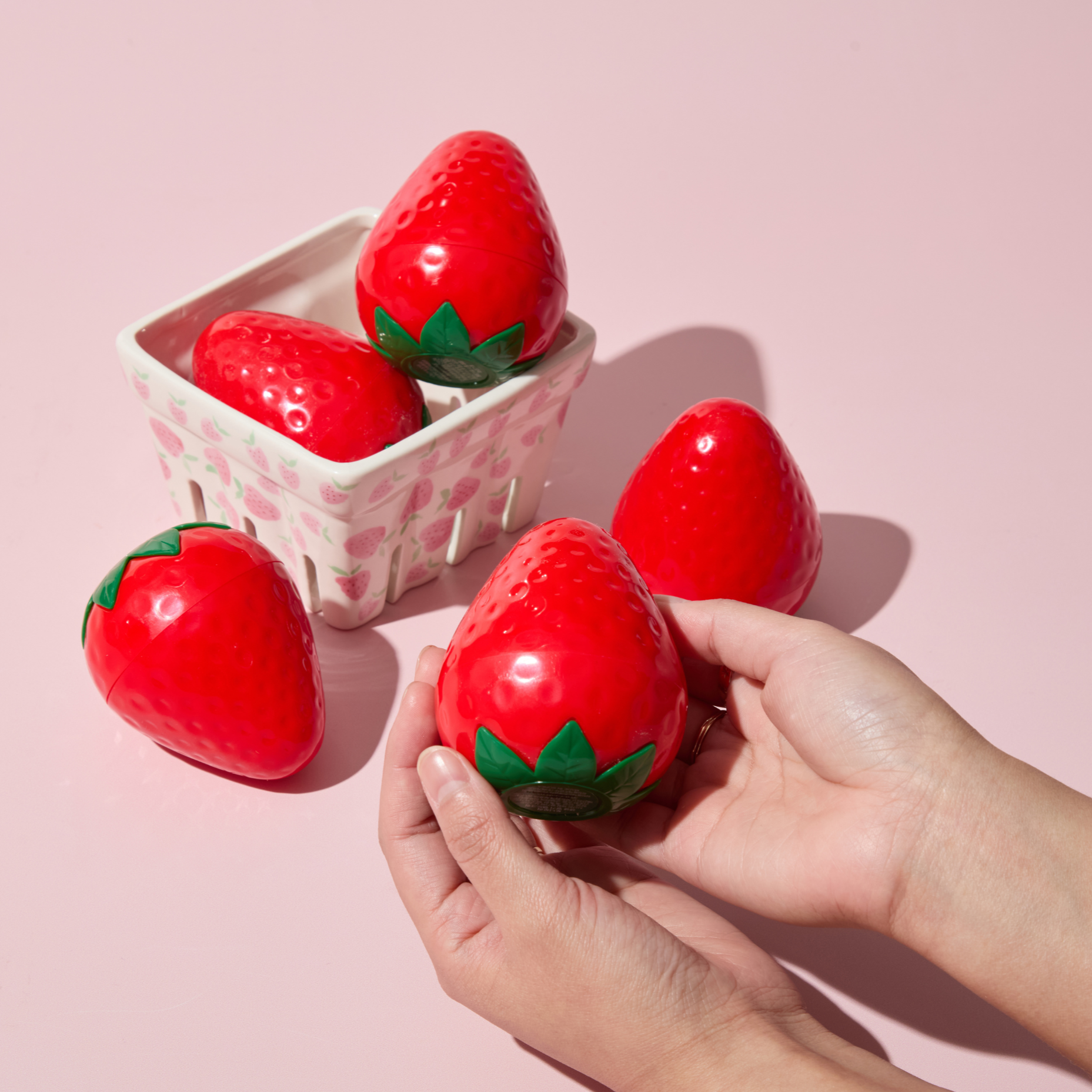 Red strawberry-shaped objects with green leaves held by a hand on a pink background