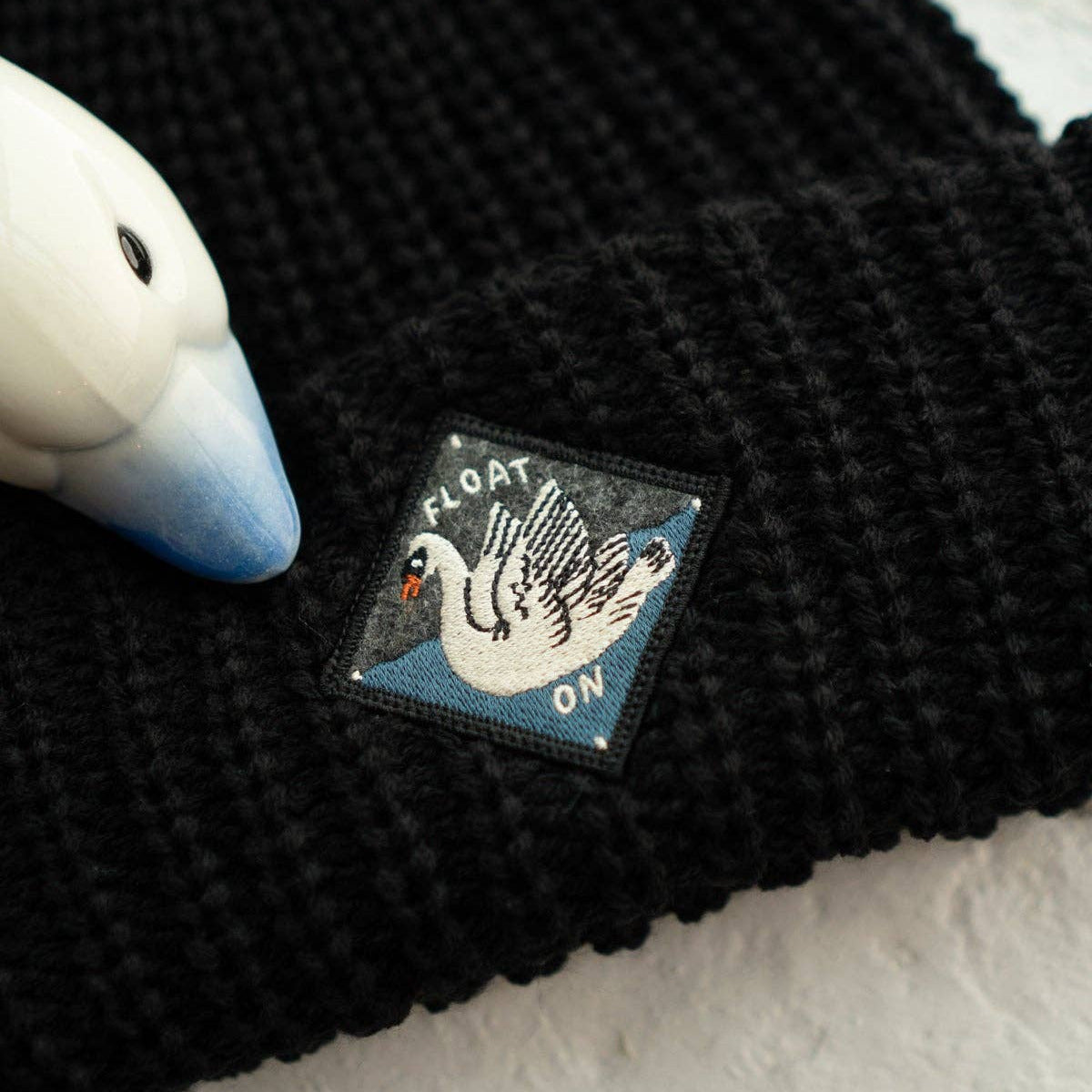 Black textured fabric with a patch featuring a swan and text on a light gray background