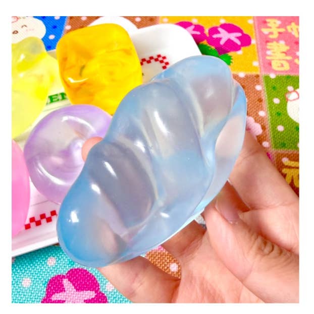 A hand holding a translucent light blue silicone ear-shaped toy, with other colorful ear-shaped toys and a patterned surface in the background.