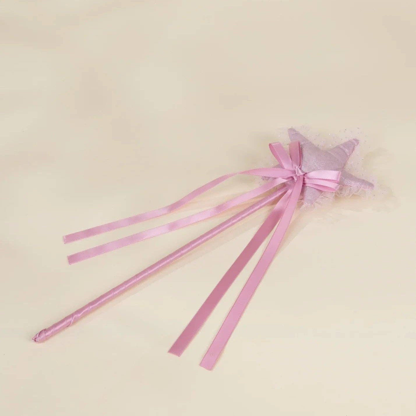 A pink star-shaped wand with long satin ribbons and a small tulle bow, photographed on a plain light beige background.