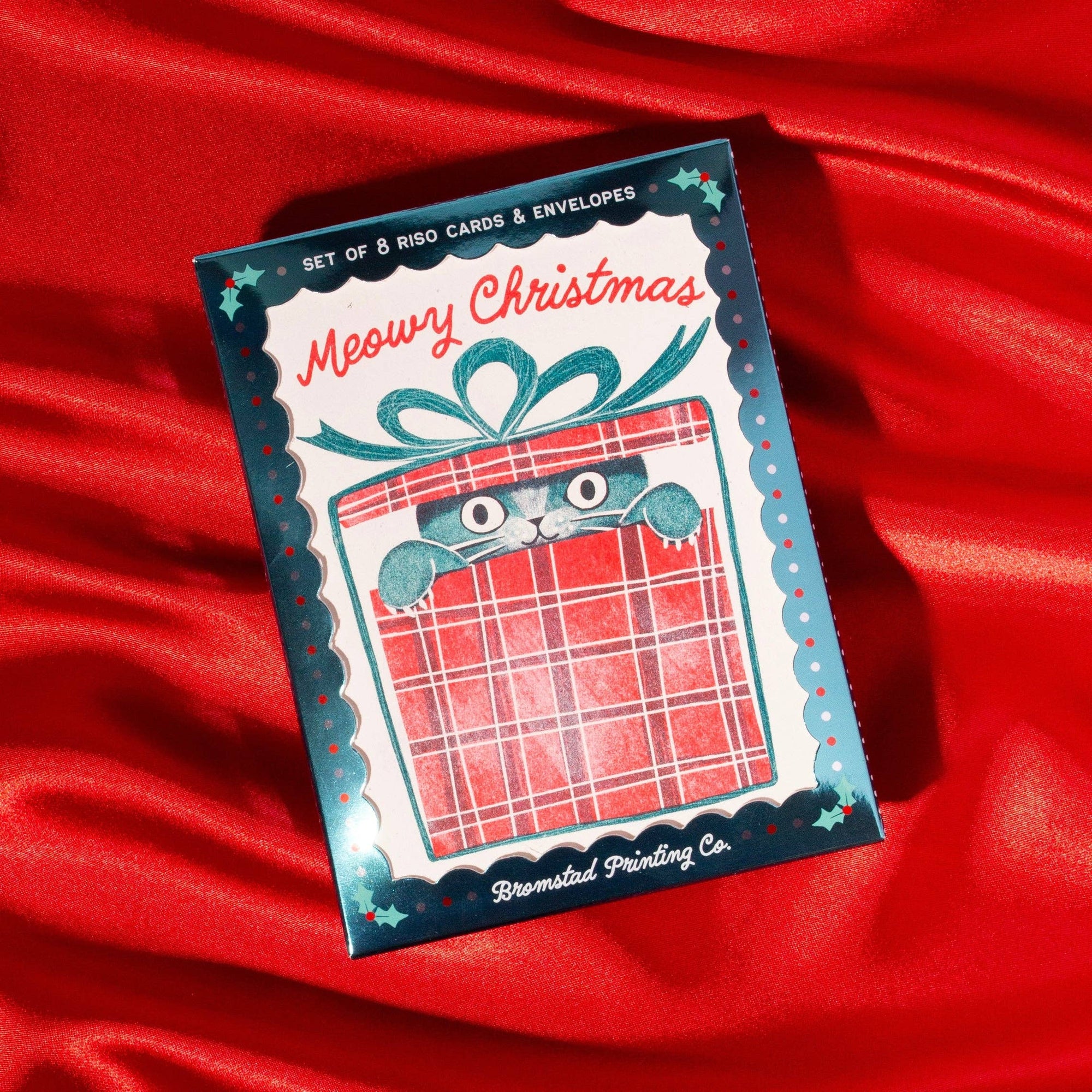 A boxed card set titled "Meowy Christmas" by Breastful Printing Co., featuring a festive illustration of a cat peeking from a wrapped gift on a red plaid background, placed on draped red fabric.