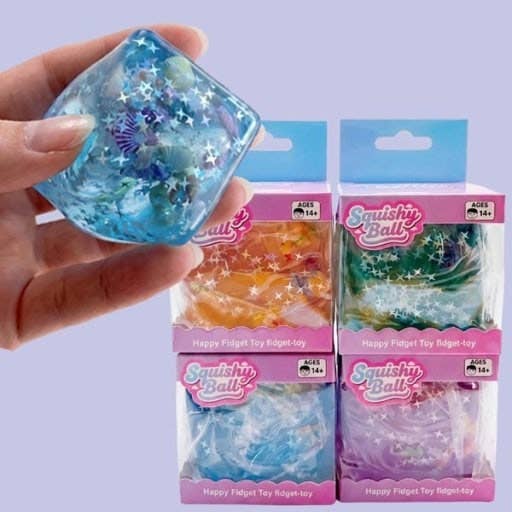 A hand holds a translucent blue squishy ball toy with star and shell details, with four packaged 'Squishy Balls' toys in orange, green, blue, and purple shown behind it on a light lavender background.
