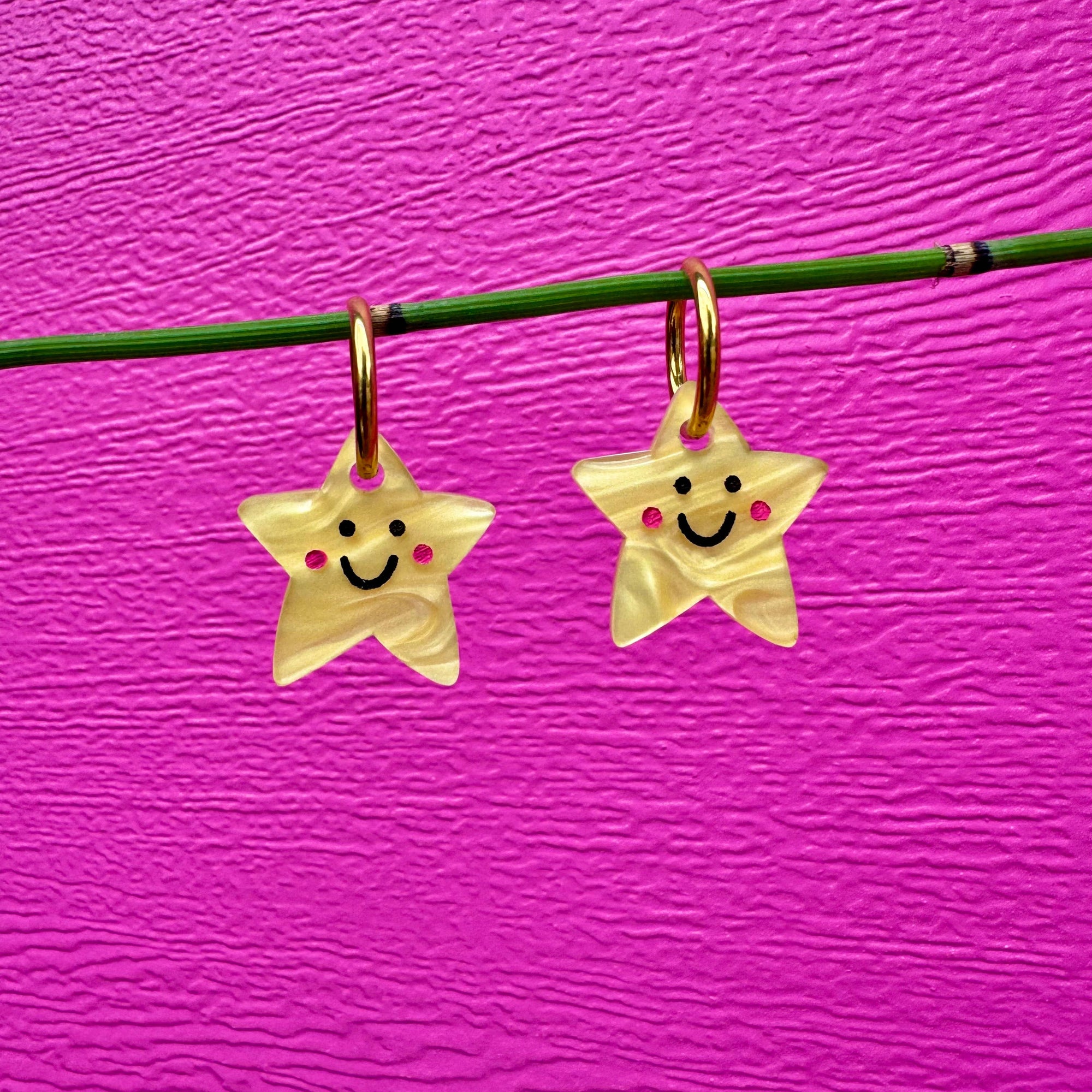 Two star-shaped earrings with smiley faces on a pink background