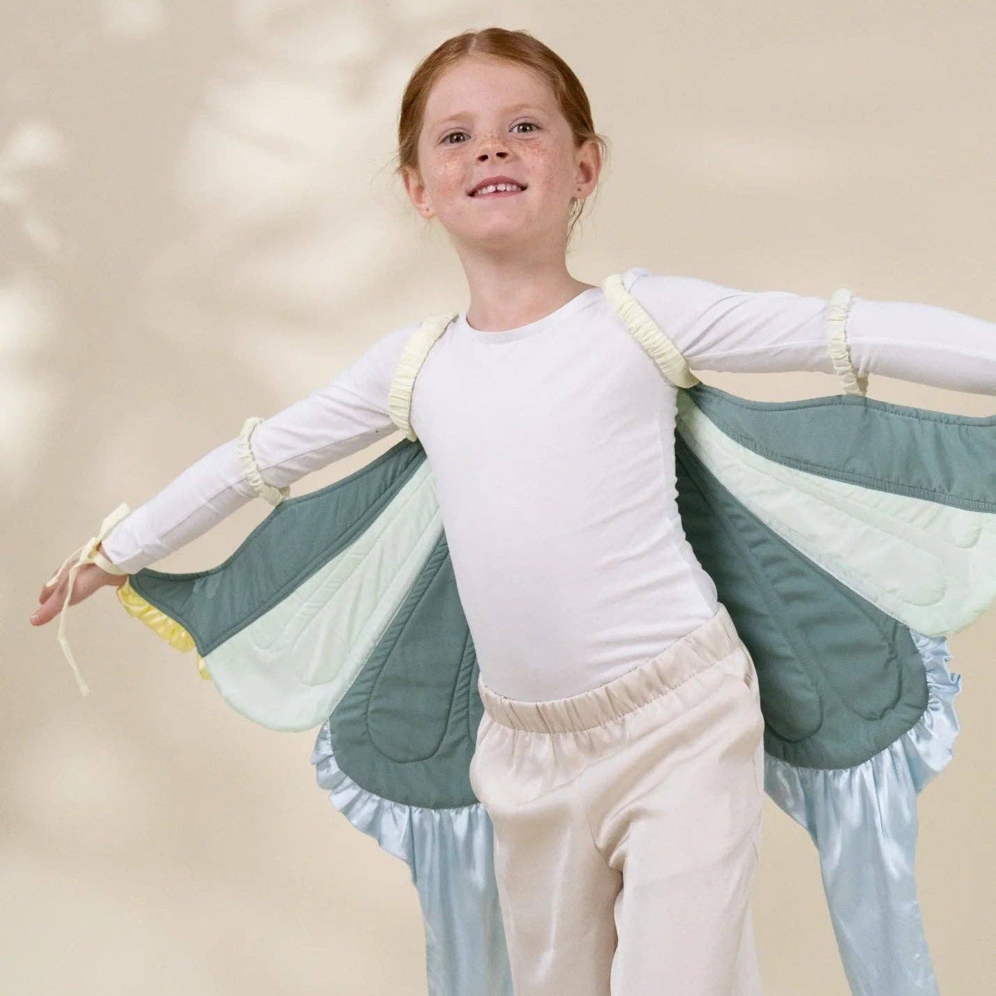 A young girl with red hair is wearing a fairy costume, featuring a white long-sleeve top, cream pants, and large quilted wings in shades of sage green, pale yellow, and light blue. She is posing with her arms outstretched against a warm beige background with soft leaf shadows.