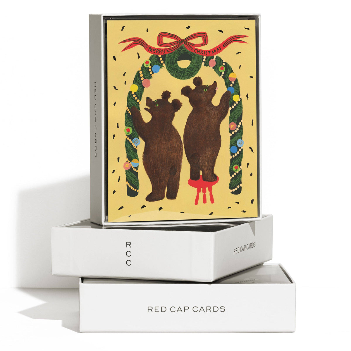 A boxed greeting card set labeled 'RED CAP CARDS' with a festive card featuring two brown bears under a green wreath and red bow on a pale yellow background, displayed on white boxes against a white studio background.