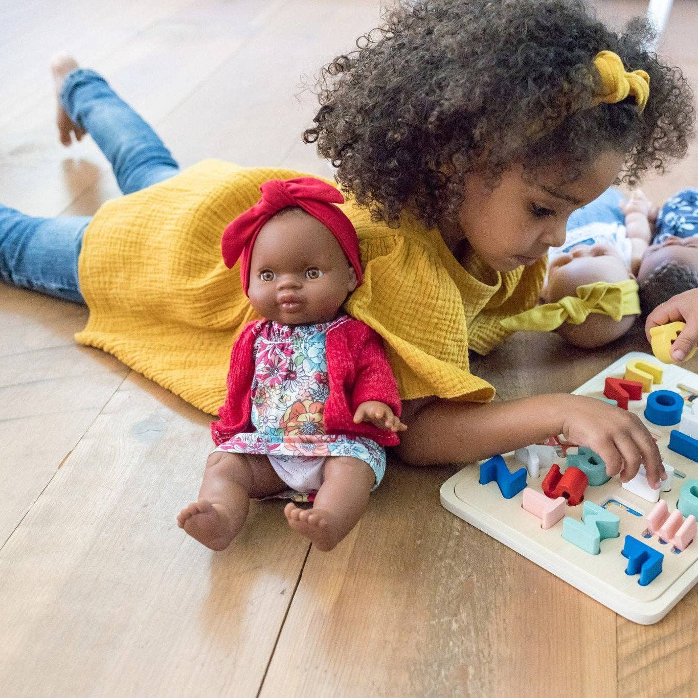 Two children playing with a doll and a puzzle on a wooden floor.
