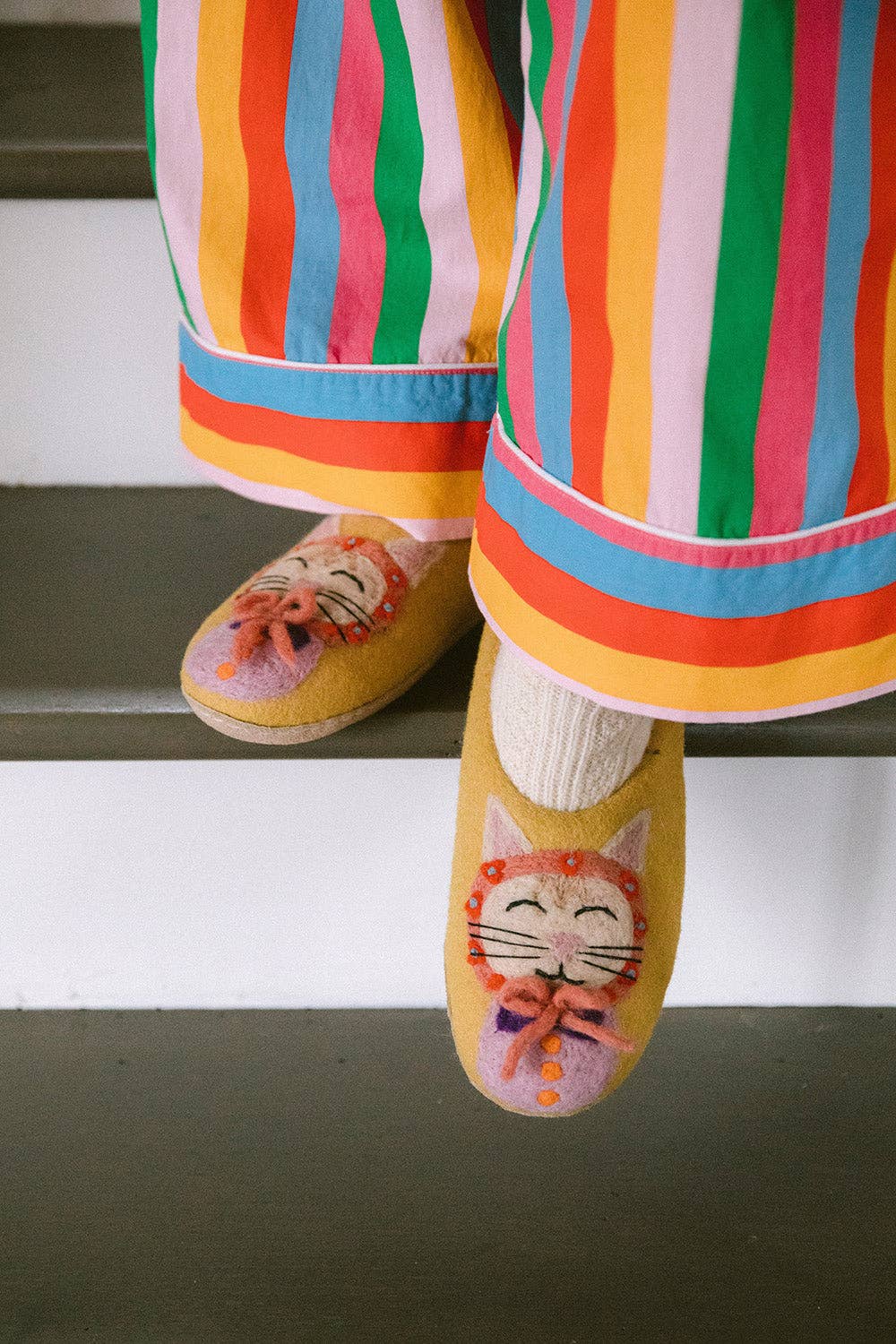 A person wearing colorful striped pajama pants and yellow slip-on slippers with a cat face design, standing on white stairs.