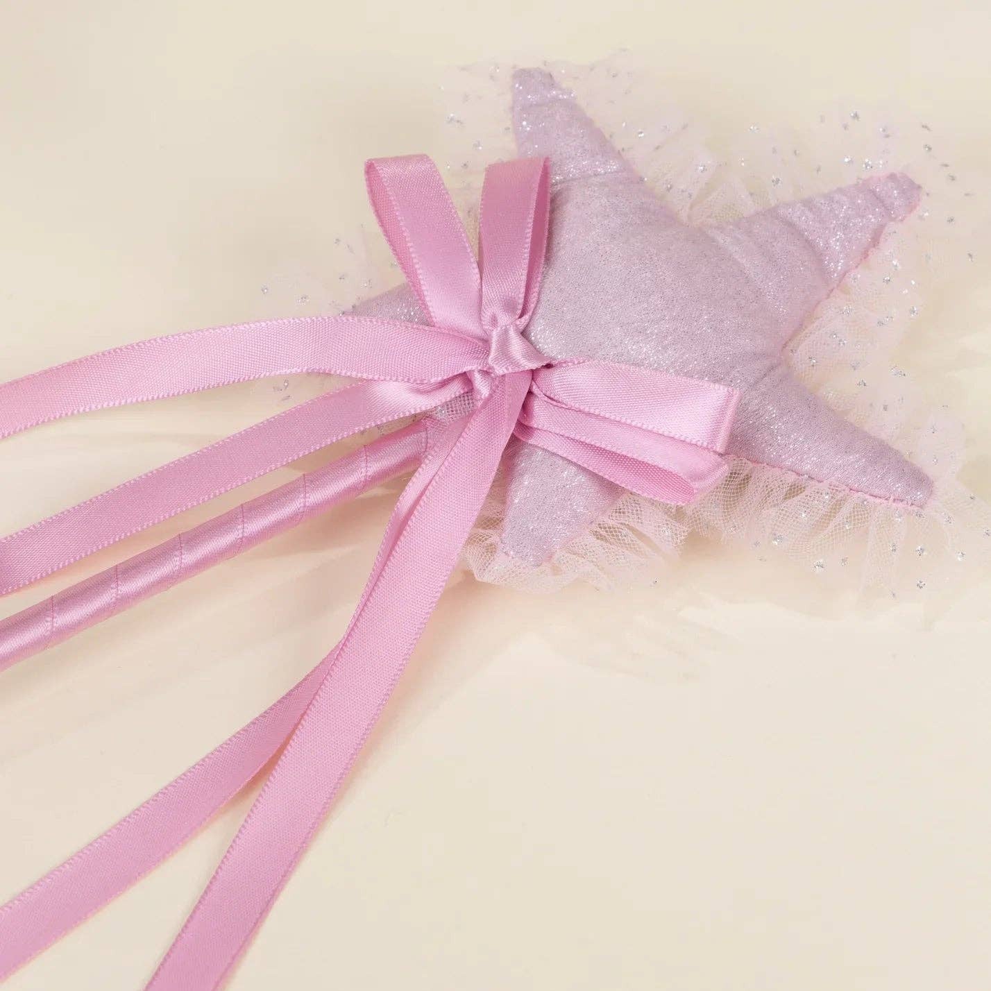 A pink fabric star wand topper with a glittery lavender star center, a pink satin bow, and long pink ribbon tails, shown on a light cream background.