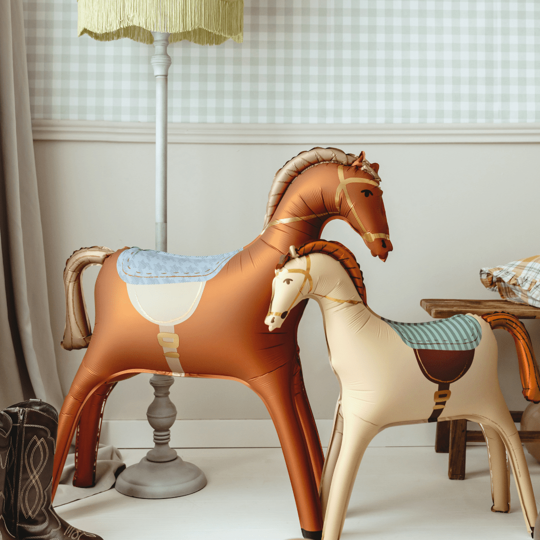 Two decorative rocking horse toys, one larger brown horse and one smaller cream-colored horse, displayed indoors next to a floor lamp and a small stool.