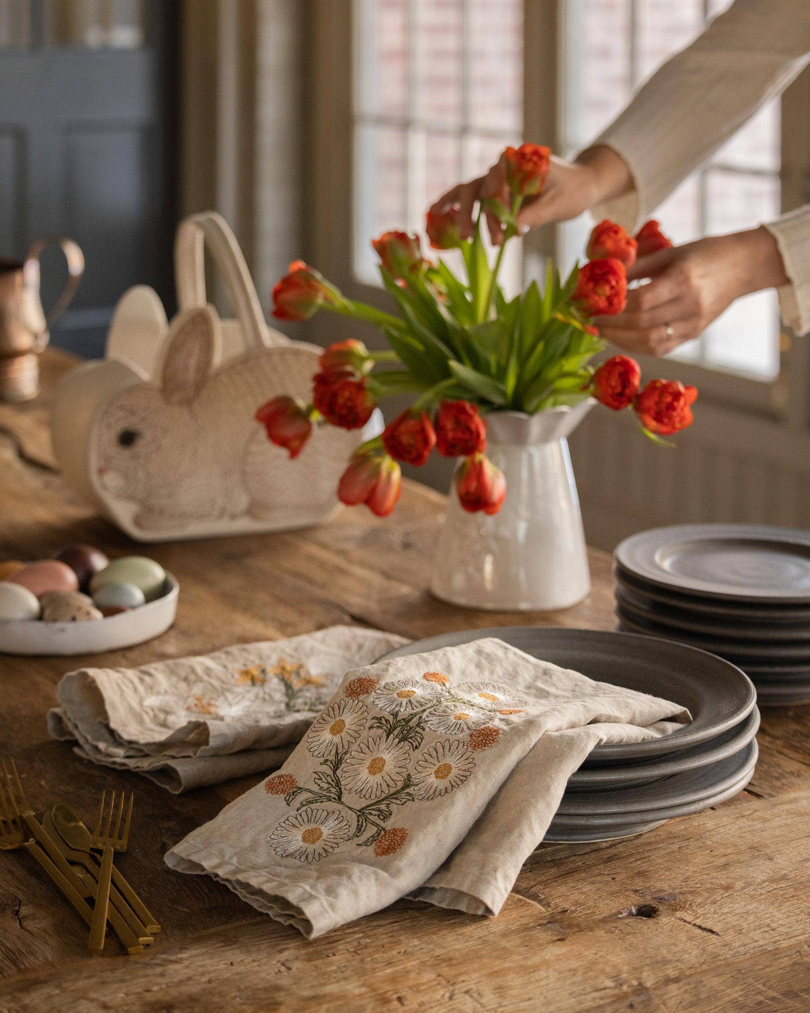 Person arranging tulips in a vase on a wooden table with Easter decorations.