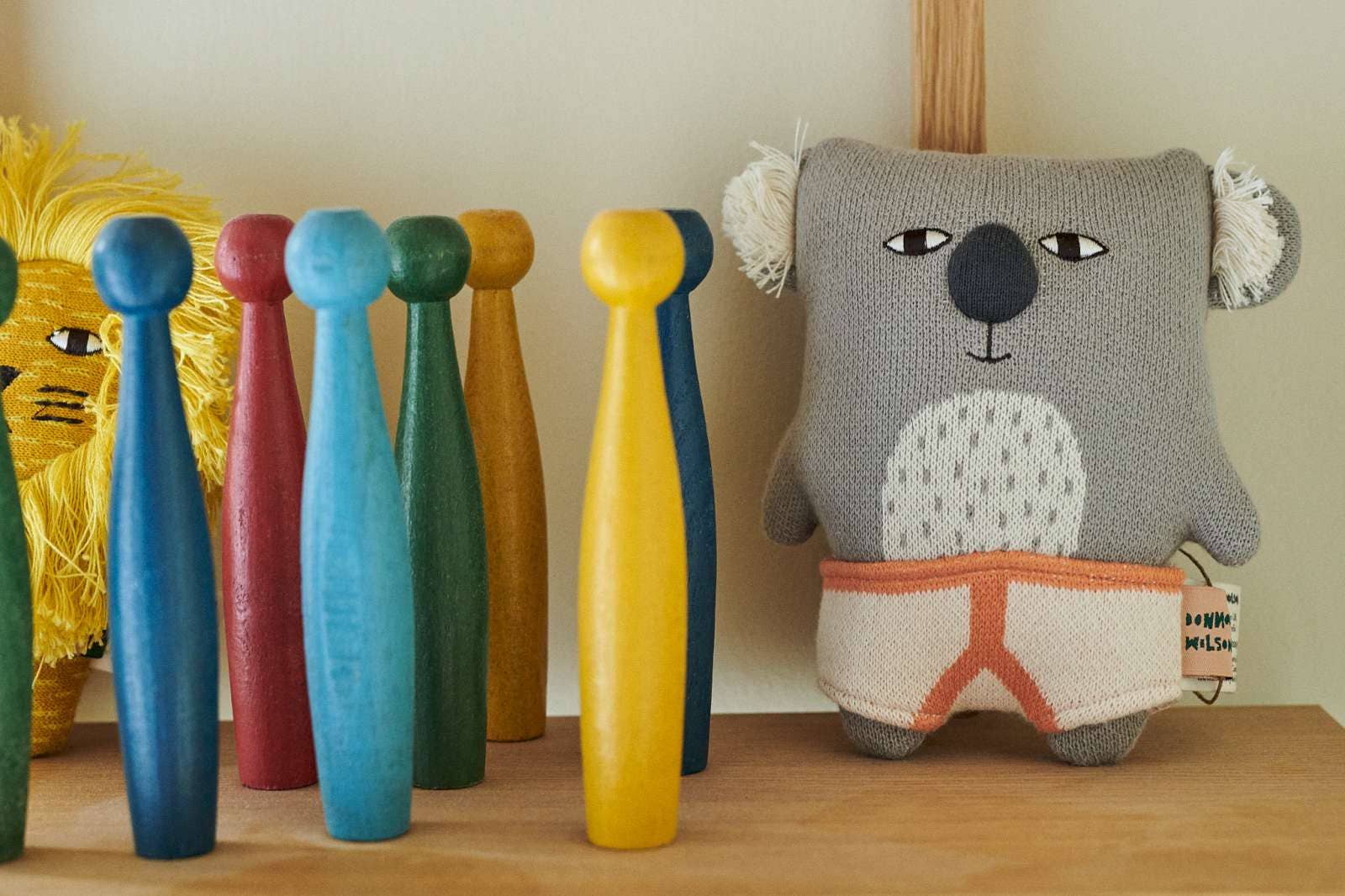 A cozy koala plush toy wearing orange shorts stands on a wooden shelf next to a row of colorful wooden toy bats. The plush toy is made of soft gray knit fabric with white ear fluffs and a white chest patch, while the bats are painted in bright shades of blue, pink, light blue, green, yellow, and dark blue.