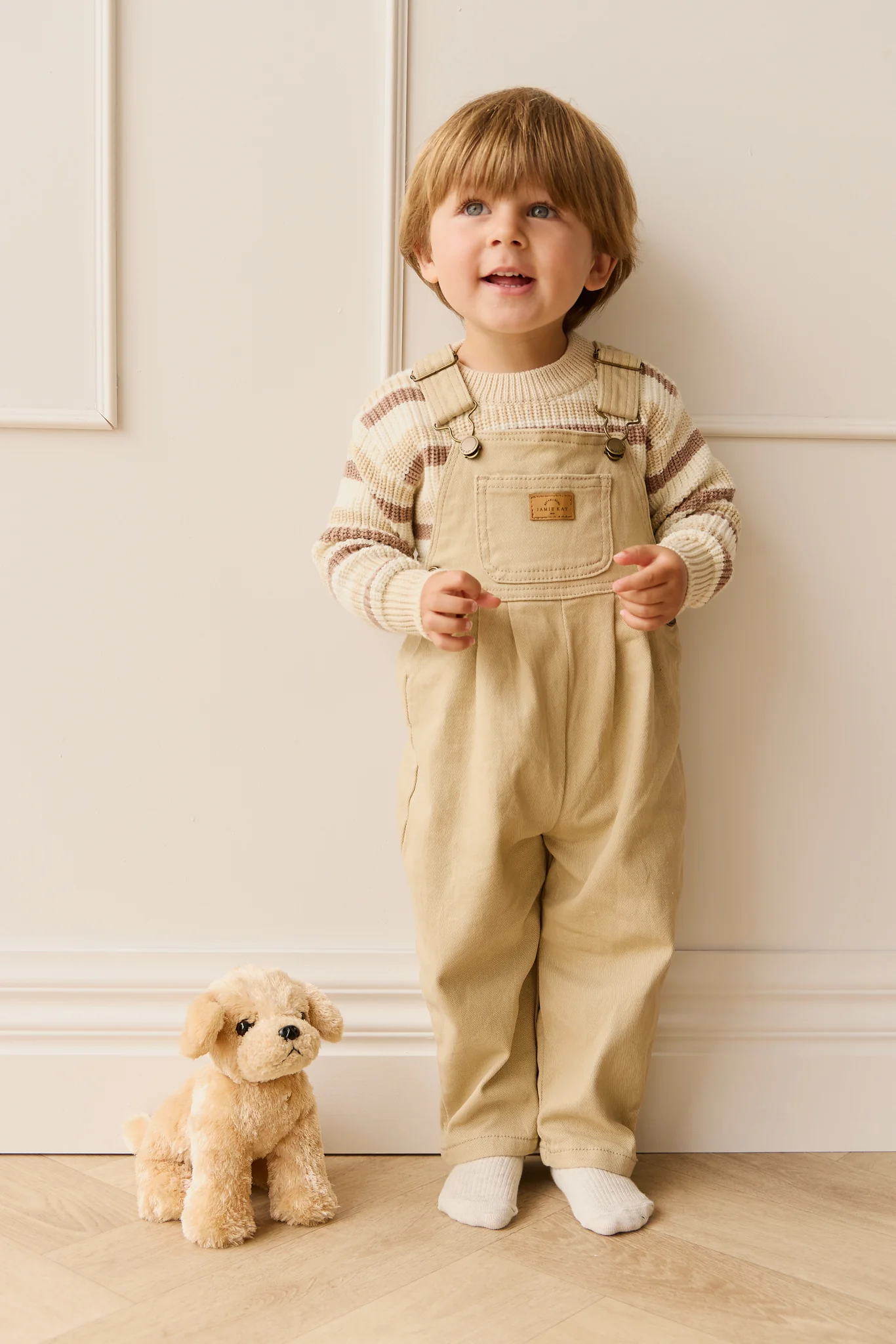 Child wearing beige overalls and striped sweater standing next to a plush toy on a wooden floor.