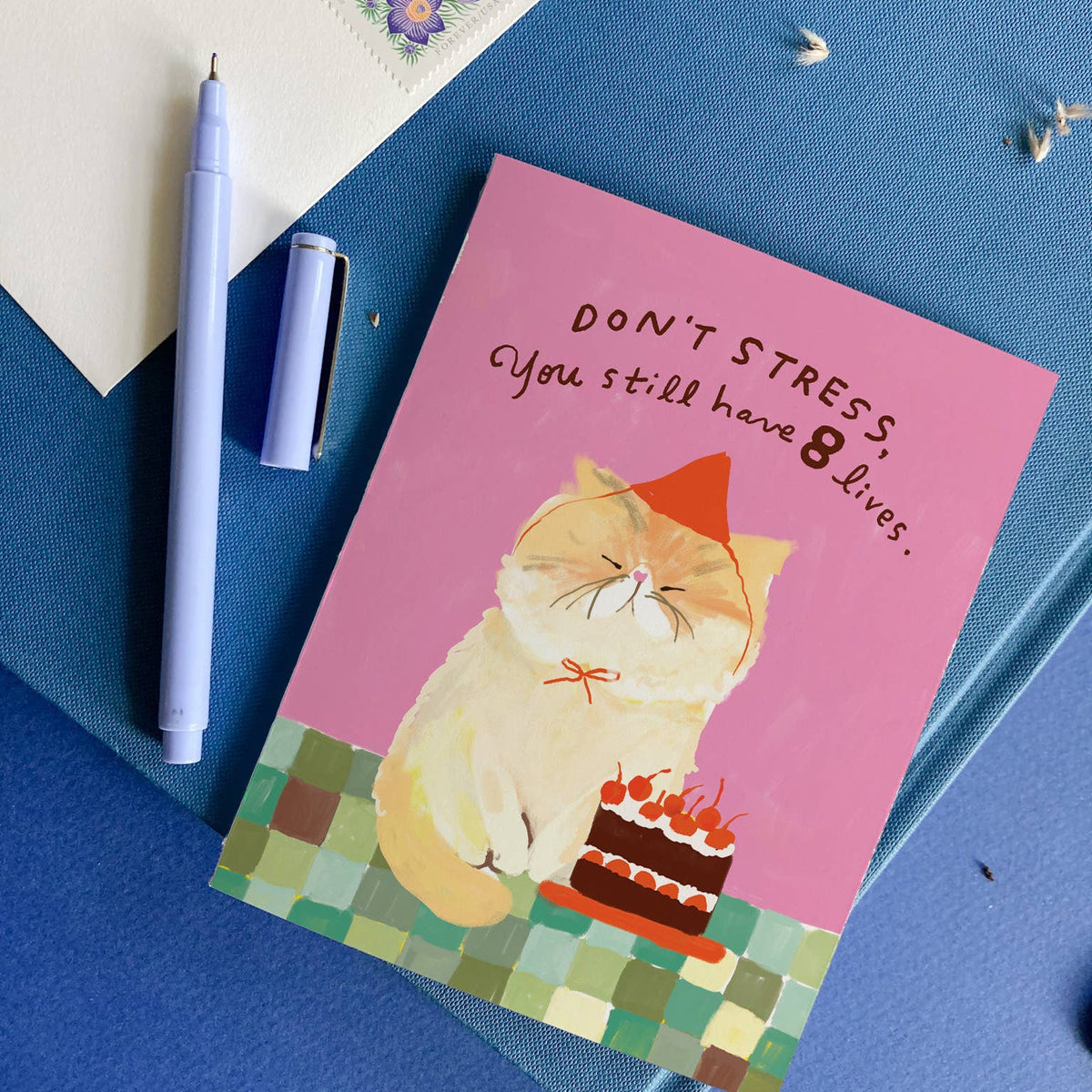 Greeting card with a cat illustration and humorous text on a blue surface with stationery items.