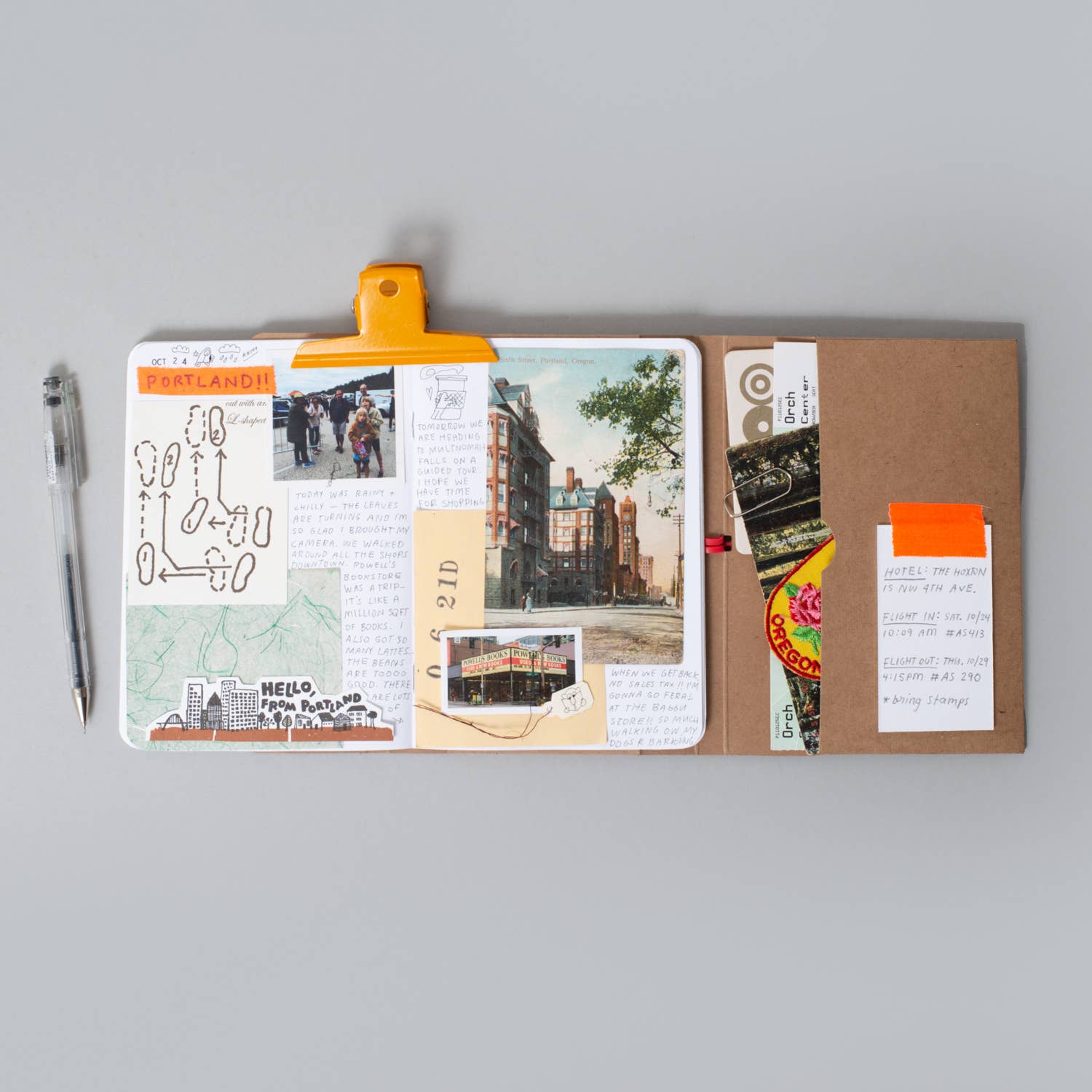 A flat-lay view of a brown kraft paper travel journal or planner cover, held open by a bright orange plastic clip. The left page is filled with travel-themed collage elements, including a map, a photo of a city street, a ticket stub, and handwritten notes.