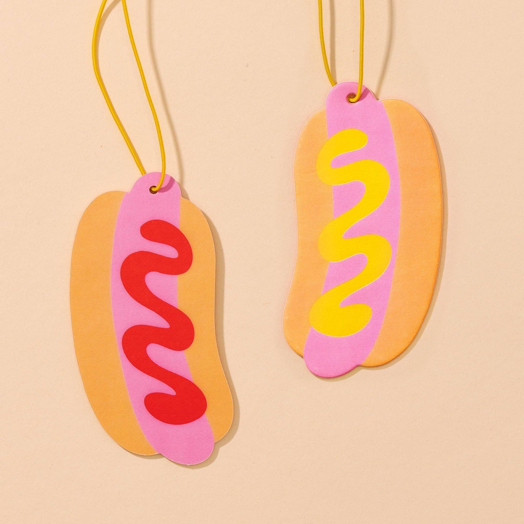 Two hot dog-shaped ornaments with pink buns, red and yellow sausages on a beige background