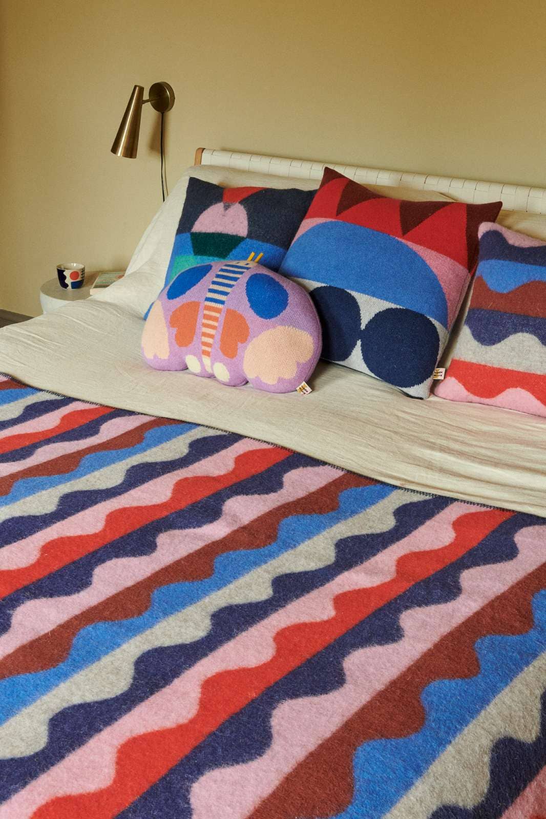 A bed styled with a colorful geometric patterned blanket, matching pillows, and a pink butterfly-shaped pillow in a warm-toned bedroom setting.